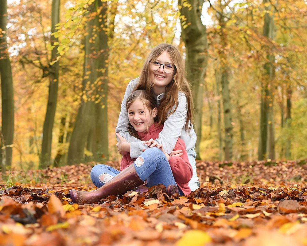 Smiling sisters playfully hold hands in an autumn forest, surrounded by golden trees and fallen leaves in a joyful scene.