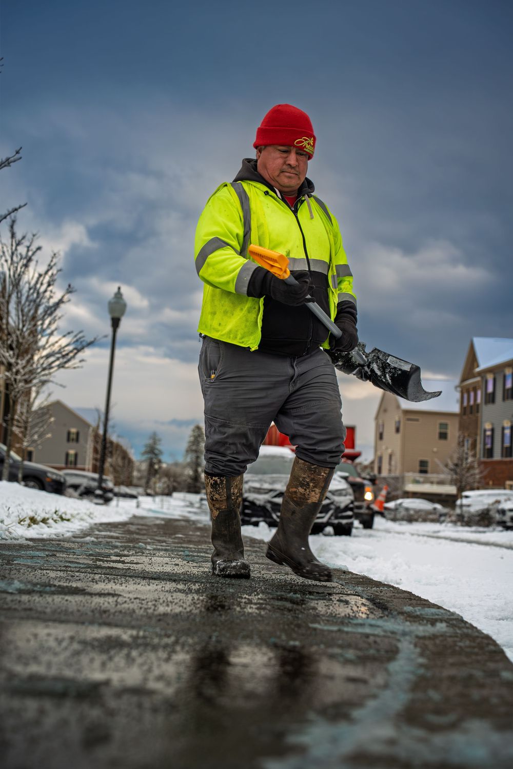 Worker in yellow hi-vis vest and red beanie shovels snow on a wet sidewalk during winter in a residential neighborhood.