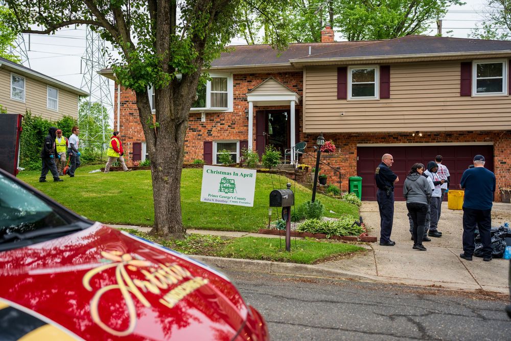 Landscaping crew and community members gather outside a brick suburban home near a Prince Georges County program sign.
