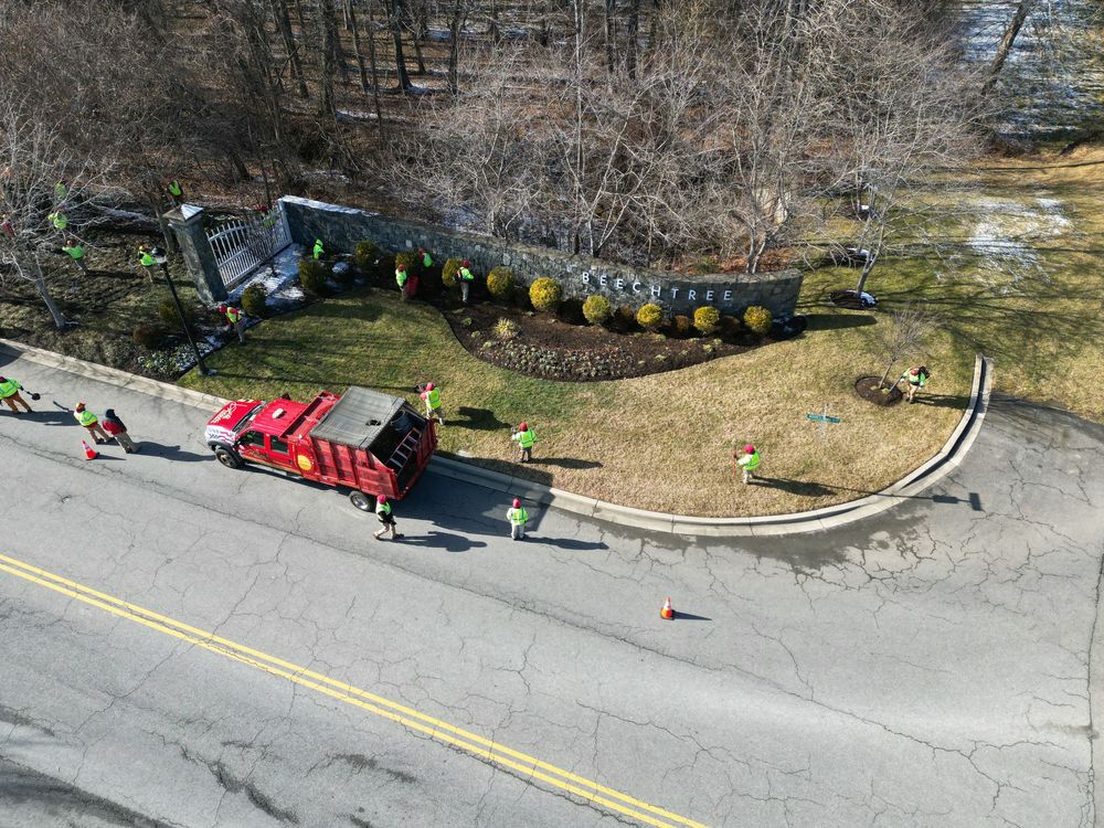 Aerial view of red trucks and workers in safety vests performing landscaping work along a curved roadside garden bed.