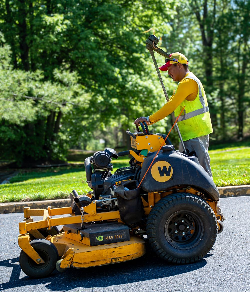 Landscaping worker in yellow safety vest operates a large zero-turn mower on a paved path.