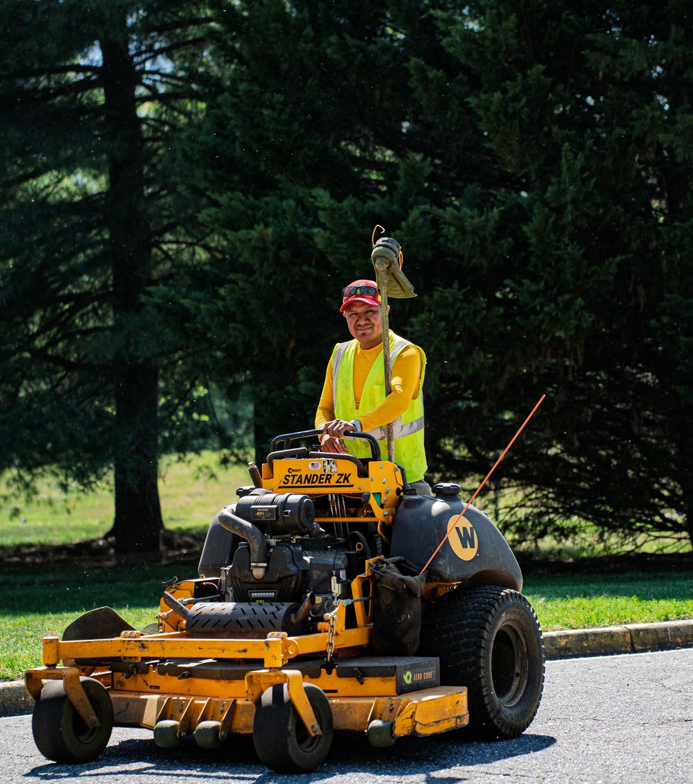 Worker in yellow safety vest operates a Wright Stander ZK stand-on mower on a paved path surrounded by green trees.