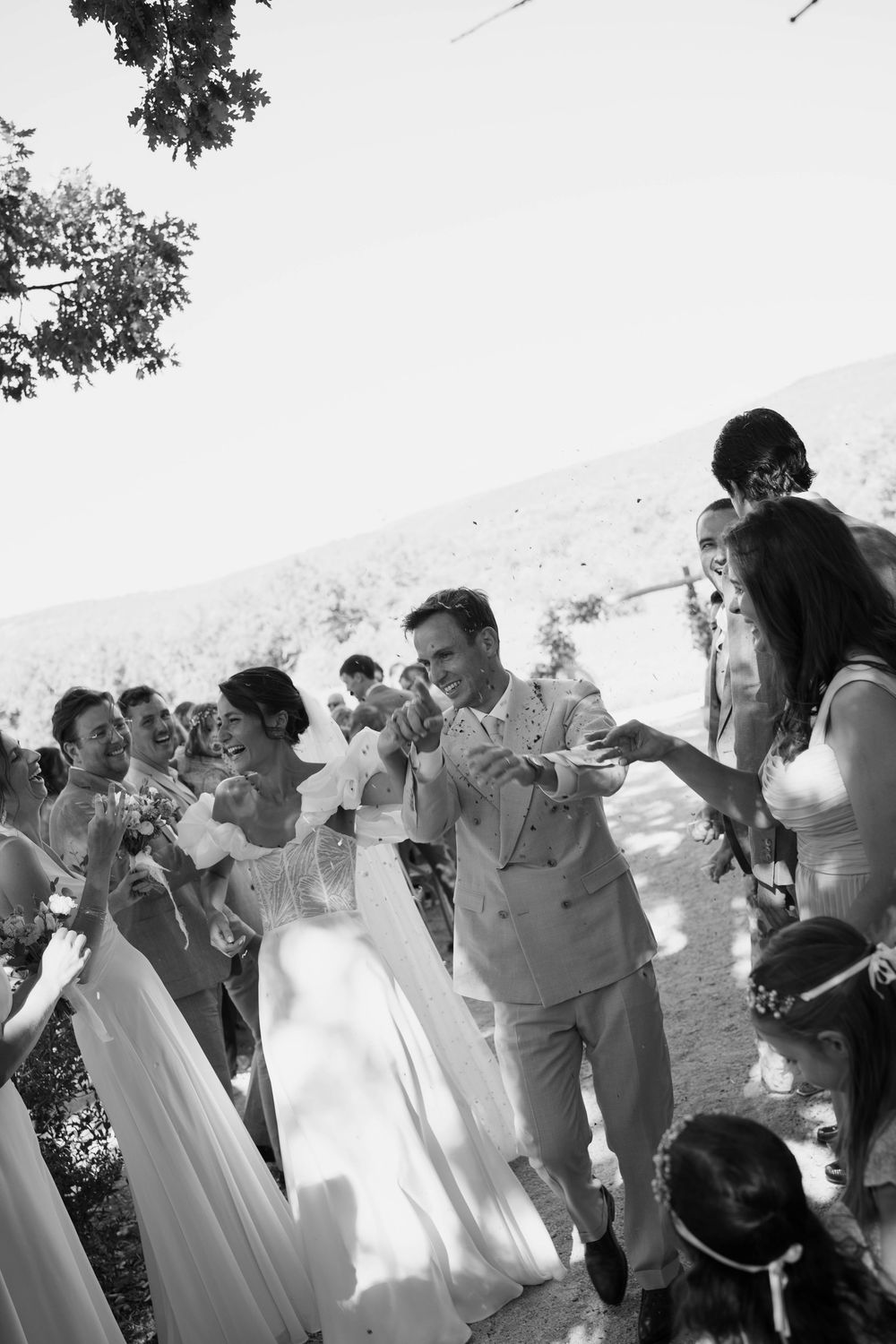 Black and white photo of a bride and groom walking through cheering guests throwing confetti at an outdoor wedding.