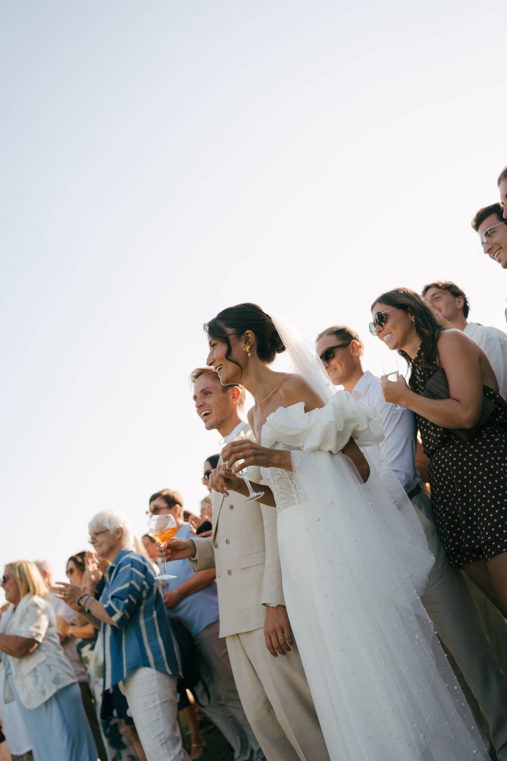La mariée en robe blanche et le marié en costume crème se tiennent parmi les invités du mariage à l'extérieur sous un ciel clair.