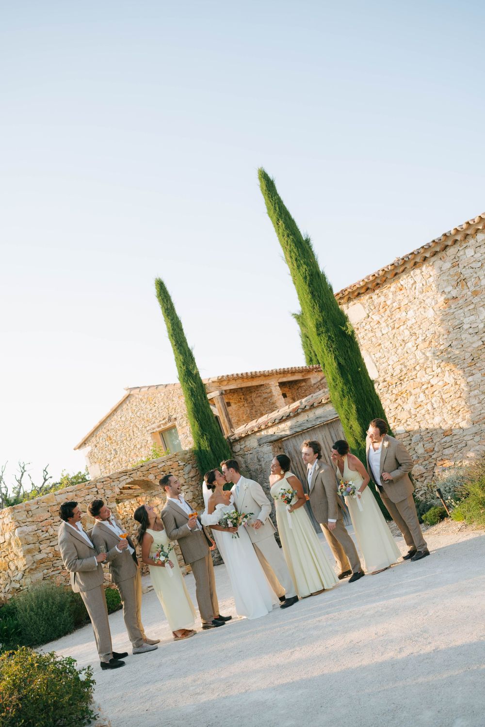 Wedding party in tan suits and white dresses poses near stone building with cypress trees in sunny outdoor setting.