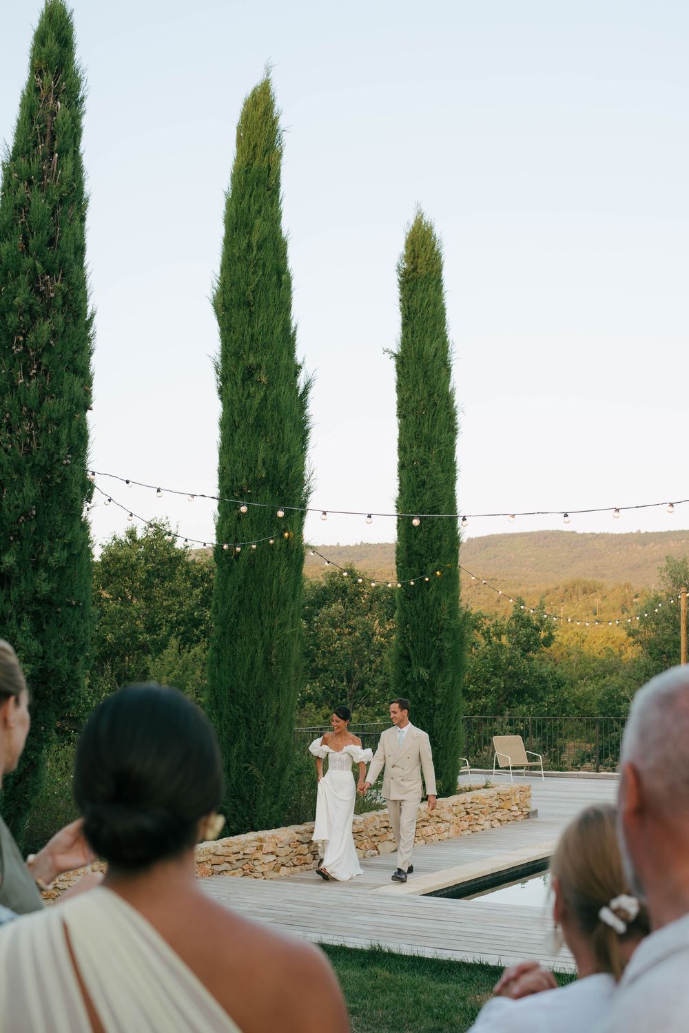 Bride and groom walk hand-in-hand at outdoor wedding reception with tall cypress trees and string lights at dusk.