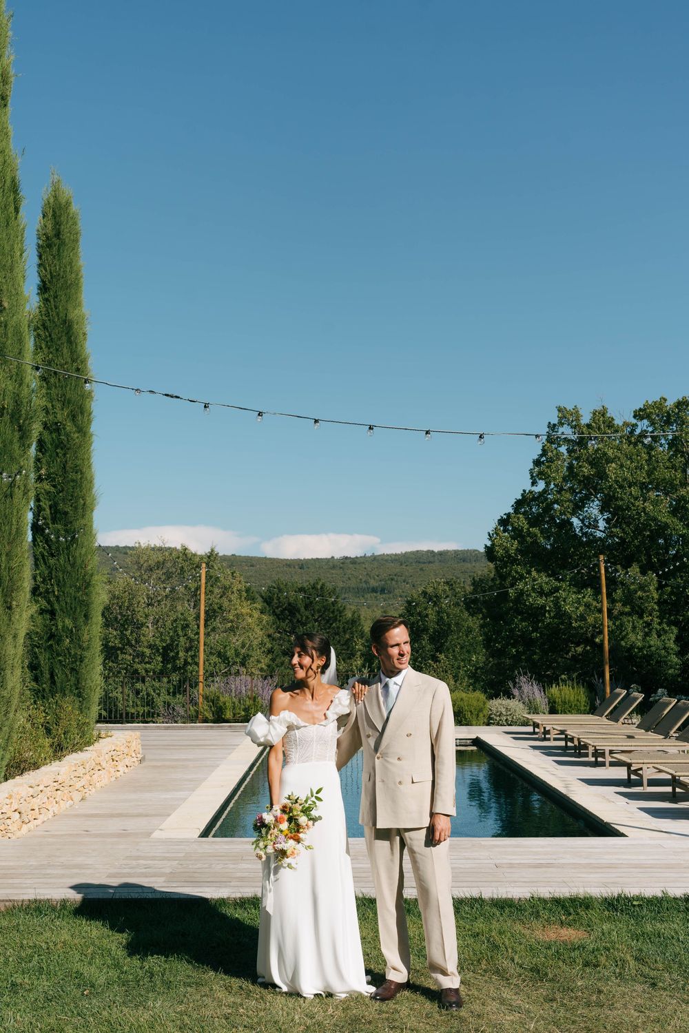 Mariée et marié posant au bord d'une piscine dans un lieu de mariage en plein air avec des cyprès et des vues sur la montagne.