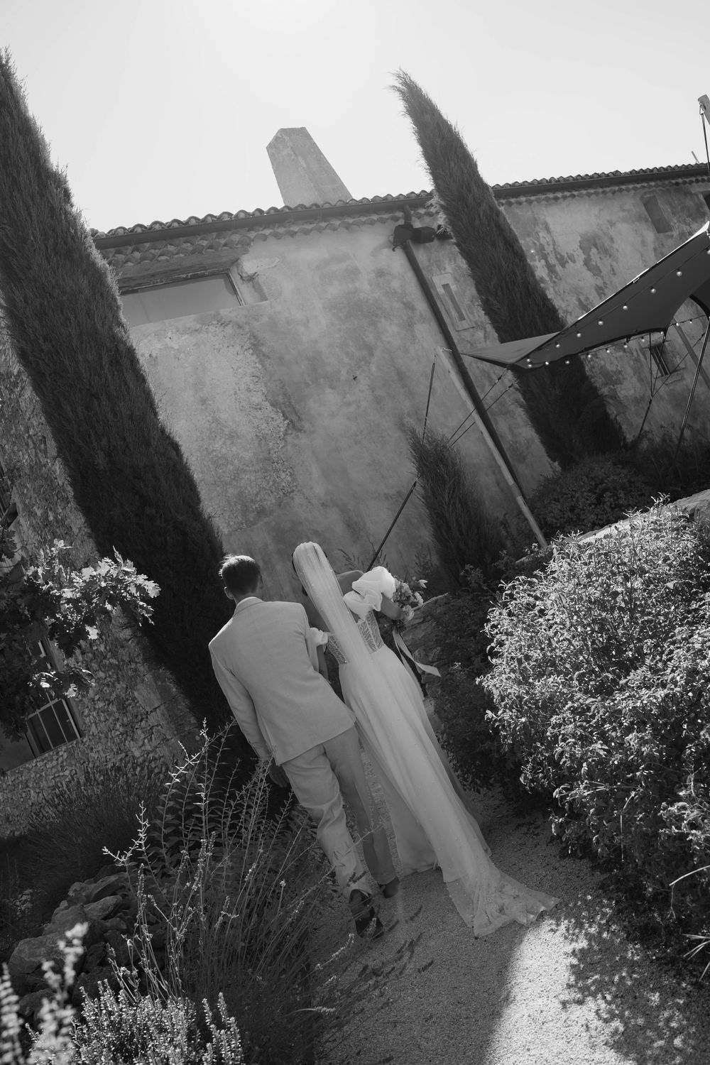 Black and white photo of a bride and groom walking hand in hand through a lush garden path beside a rustic stone building.