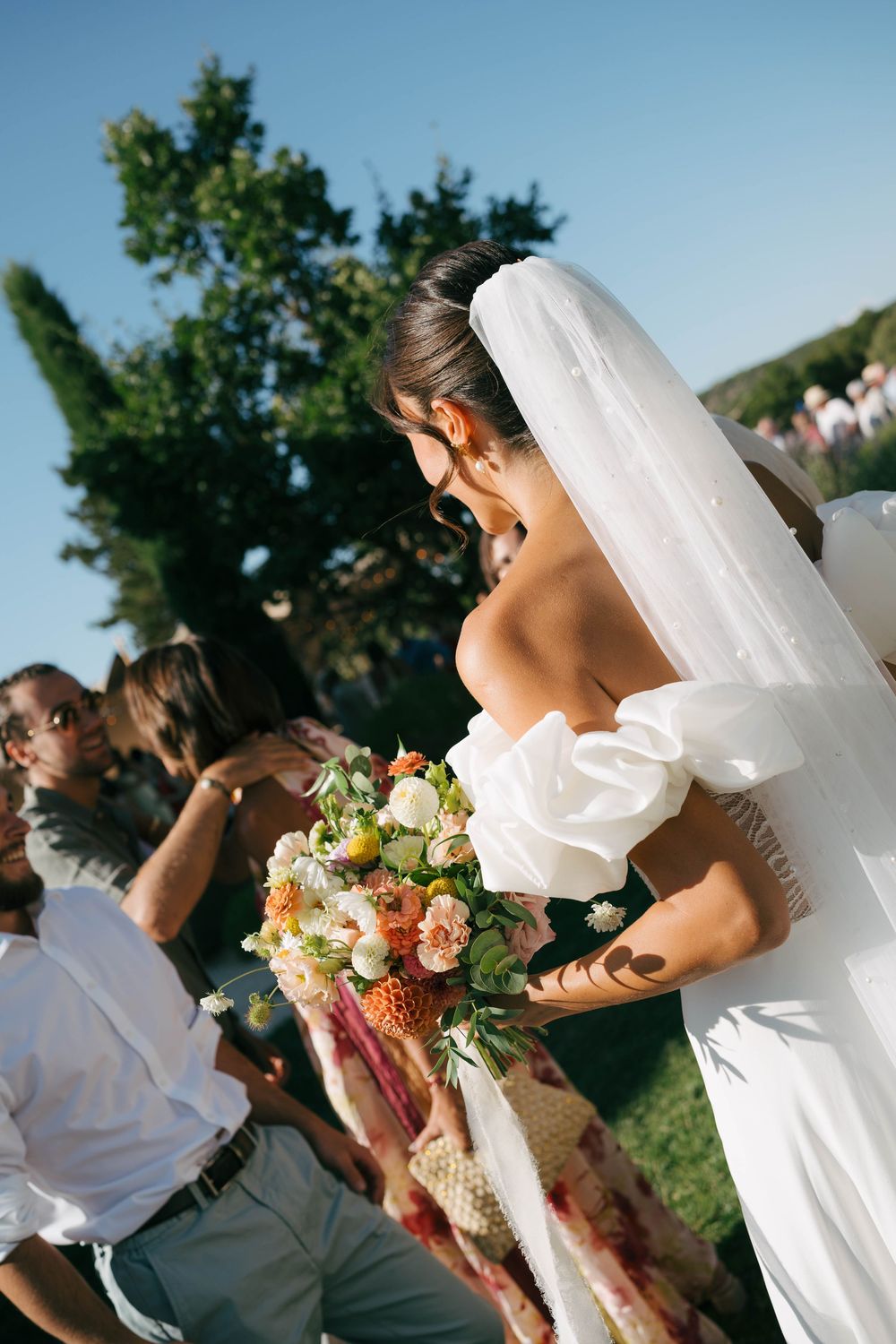 La mariée en robe blanche à épaules dénudées et au voile rit tout en tenant un bouquet coloré lors de la cérémonie de mariage en plein air.