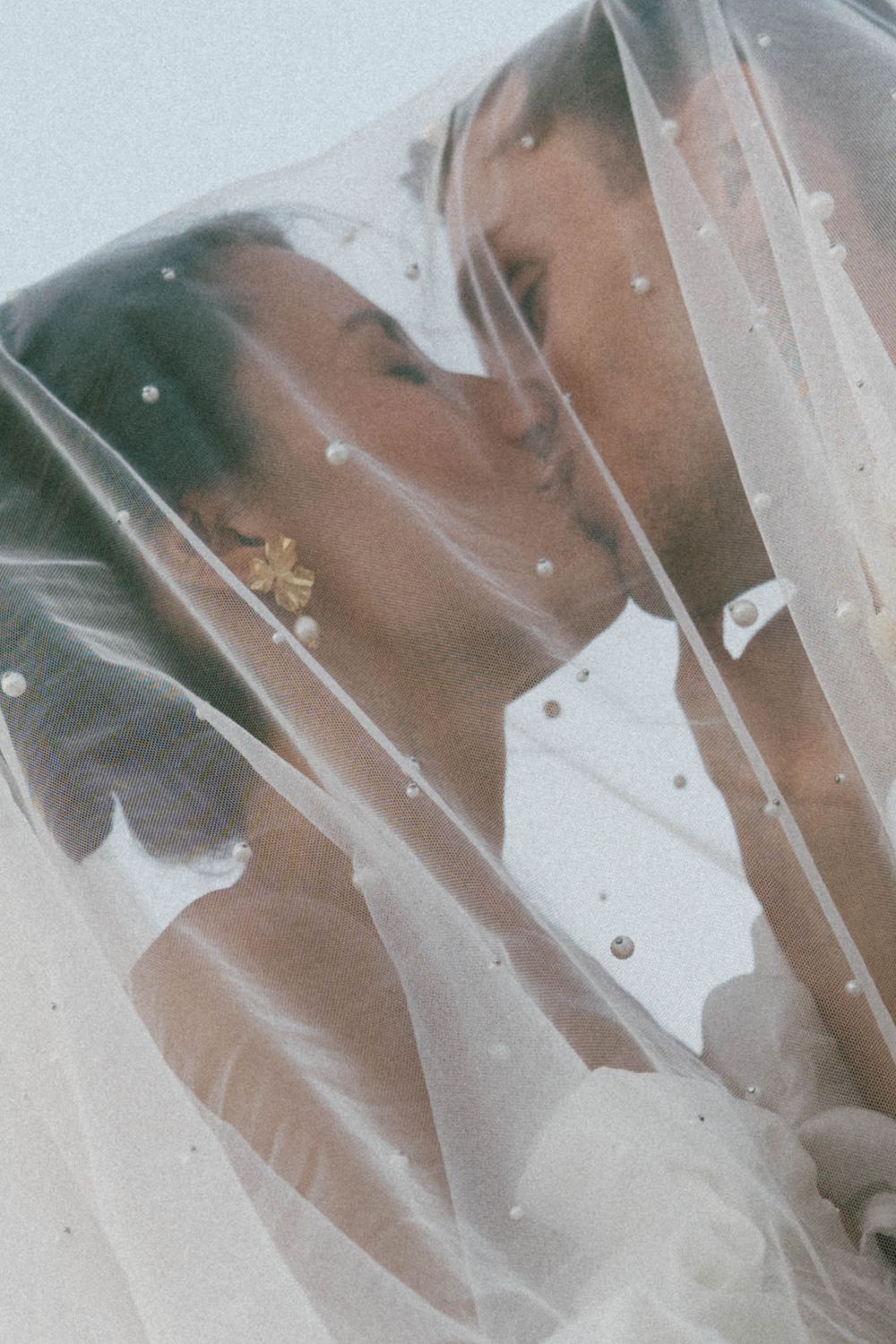 La mariée et le marié partagent un baiser romantique sous un voile de mariage orné de perles lors de leur journée spéciale.