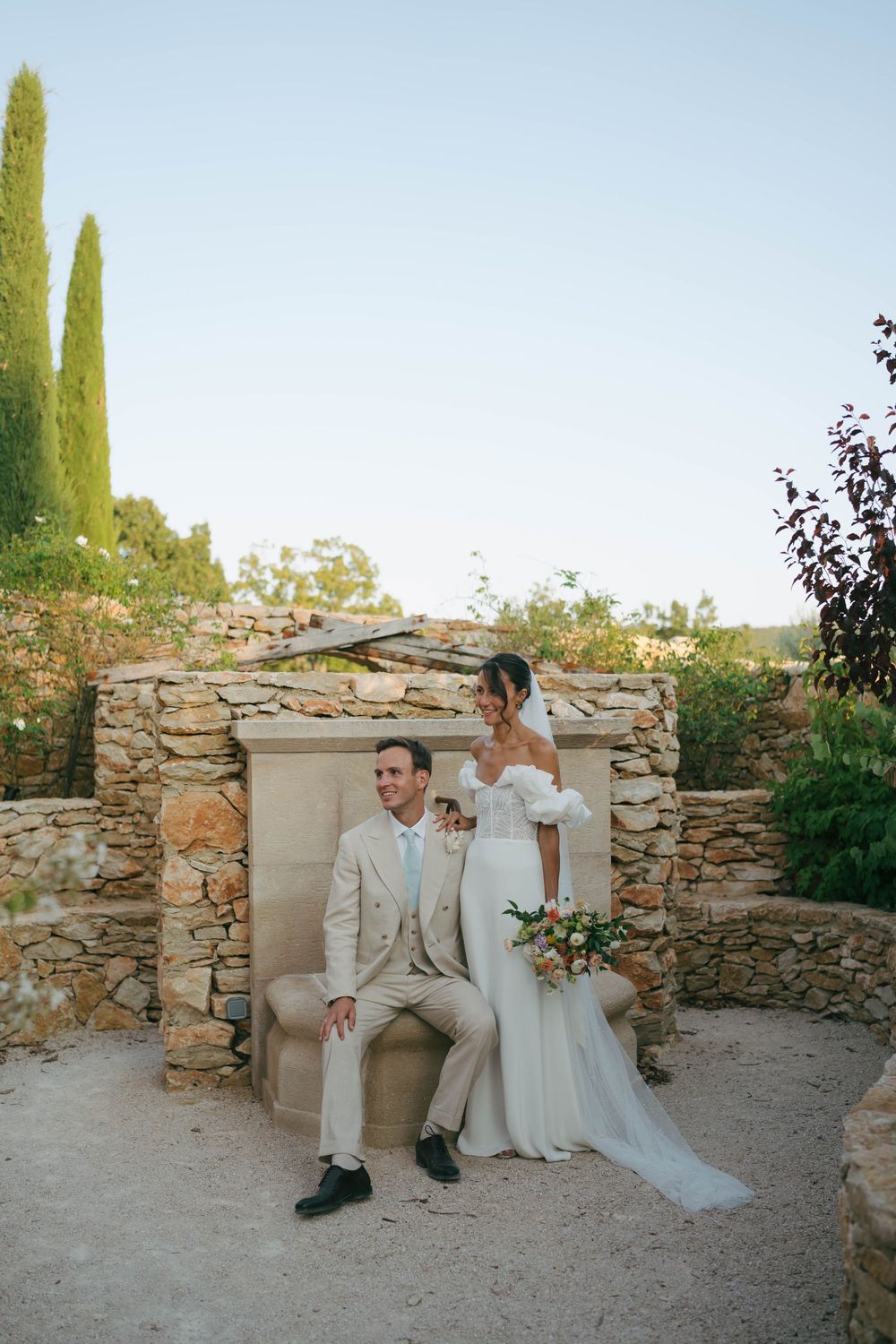 Bride in white off-shoulder gown and groom in beige suit pose by rustic stone wall at outdoor wedding venue.