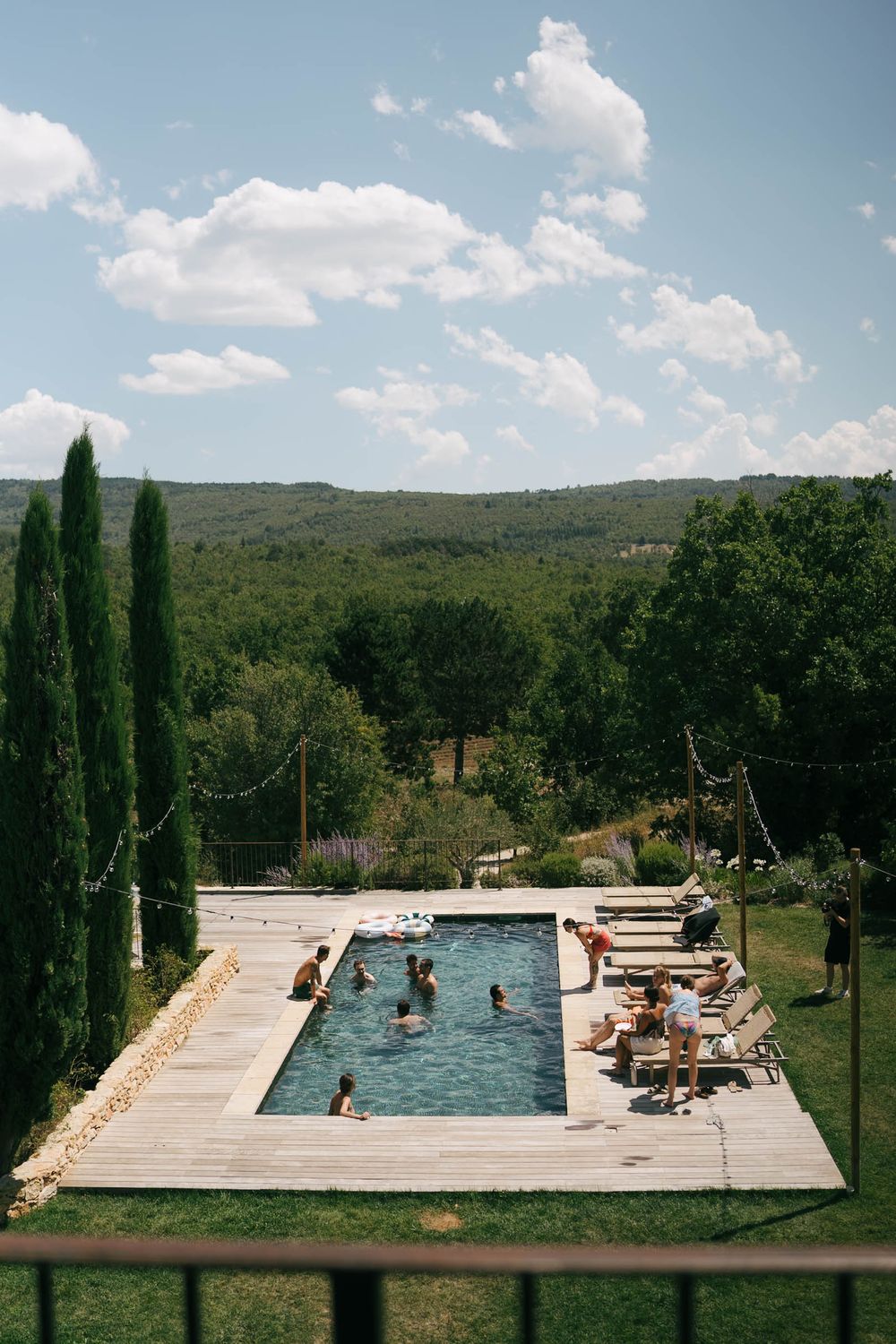 Piscine extérieure entourée de cyprès avec des nageurs, des chaises longues et des collines verdoyantes sous un ciel partiellement nuageux.