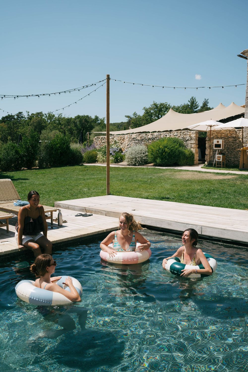 Quatre personnes profitent d'une journée ensoleillée dans une piscine avec des anneaux gonflables, entourées d'un jardin luxuriant et d'un b