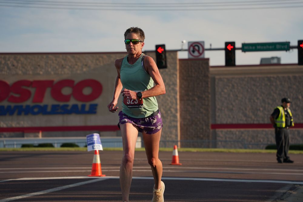 Woman running race near Costco Wholesale, wearing green tank and purple shorts, with traffic cones and officer in background.
