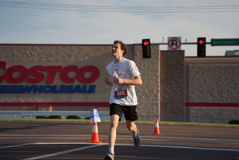 Runner wearing bib number competing in road race near Costco Wholesale store with traffic cones and traffic lights visible.