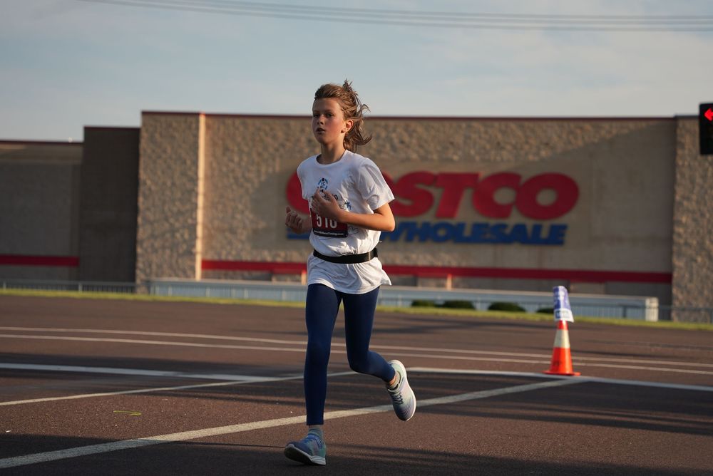 Young girl running a race near a Costco Wholesale store, wearing a race bib and athletic gear.