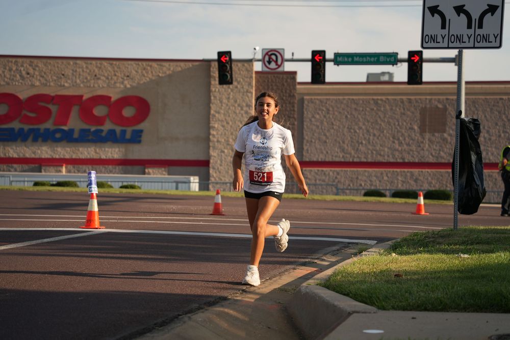 Runner in white shirt races past Costco Wholesale store at a road race event with traffic cones and signals visible.