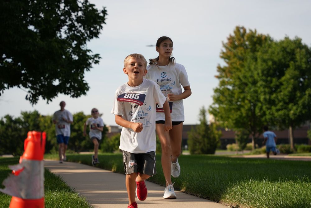 Children running in a 5K race on a suburban sidewalk, wearing race bibs, with green trees and grass in the background.