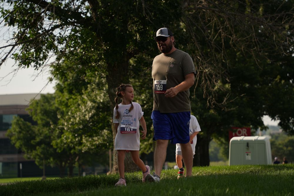 A man and young girl walking together on green grass during an outdoor community running event.