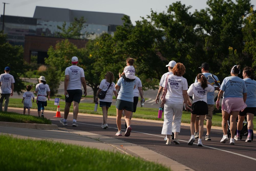 Group of participants in white t-shirts walking together during a charity fun run event on a sunny day.