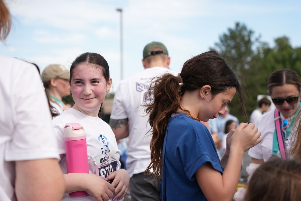 Two young girls smiling and chatting outdoors at a community fun run event on a sunny day.