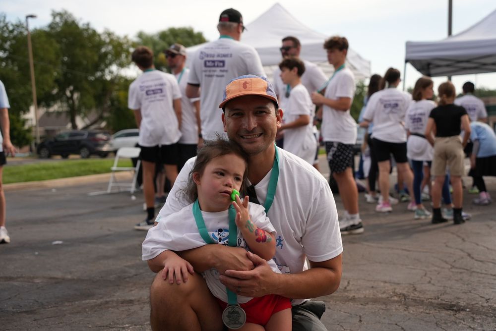 Man and young girl pose together at outdoor charity run event, both wearing white t-shirts, man displaying finisher medal.
