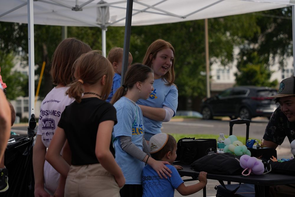 Children gather under a white tent at an outdoor community event on a sunny summer day.