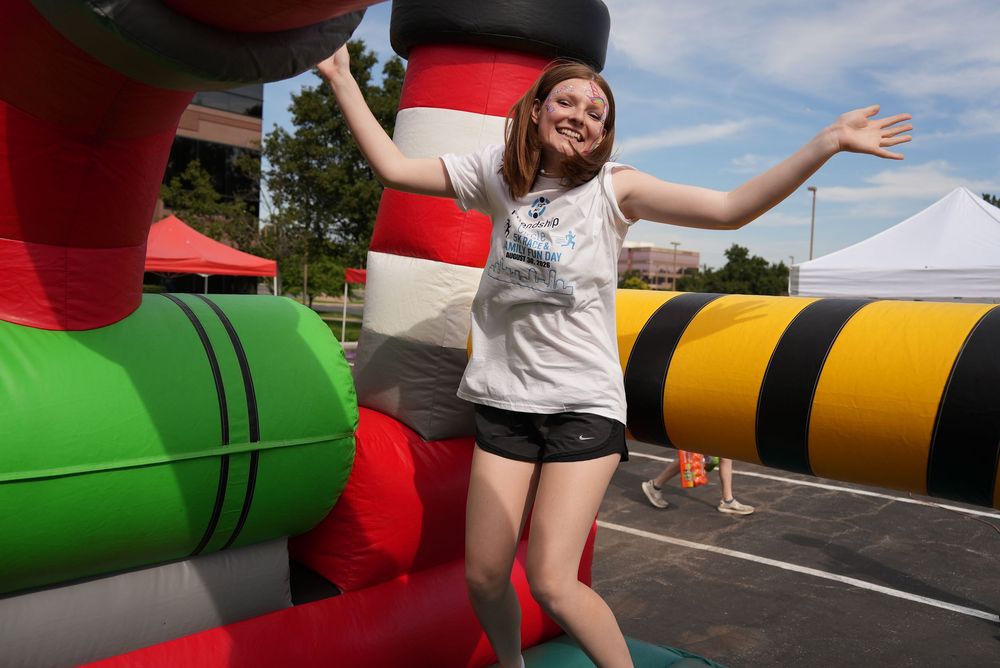 Young woman smiling with arms outstretched near colorful inflatable bounce house at outdoor carnival event.