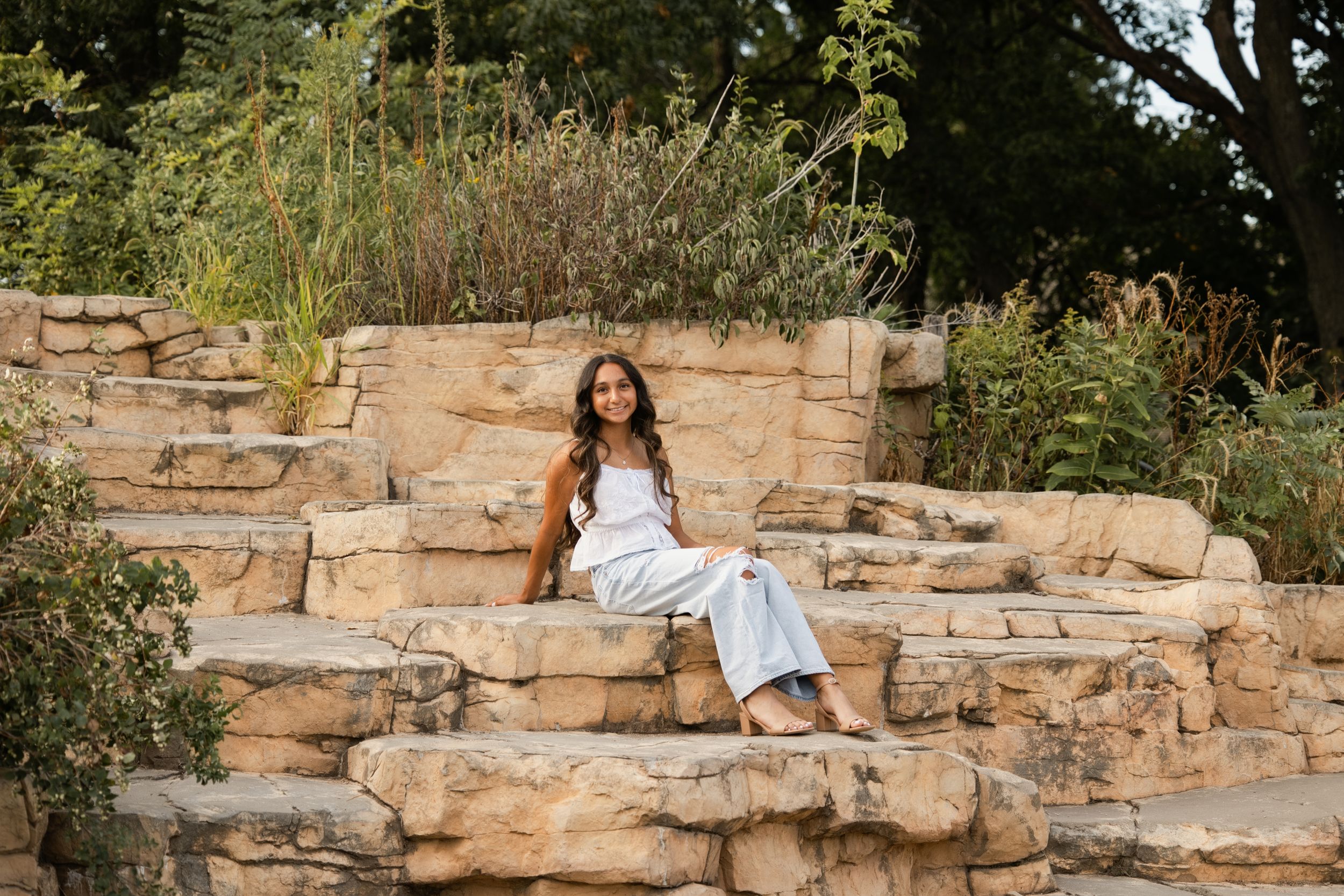 Woman in white outfit sitting on natural stone steps surrounded by lush greenery in an outdoor garden setting.