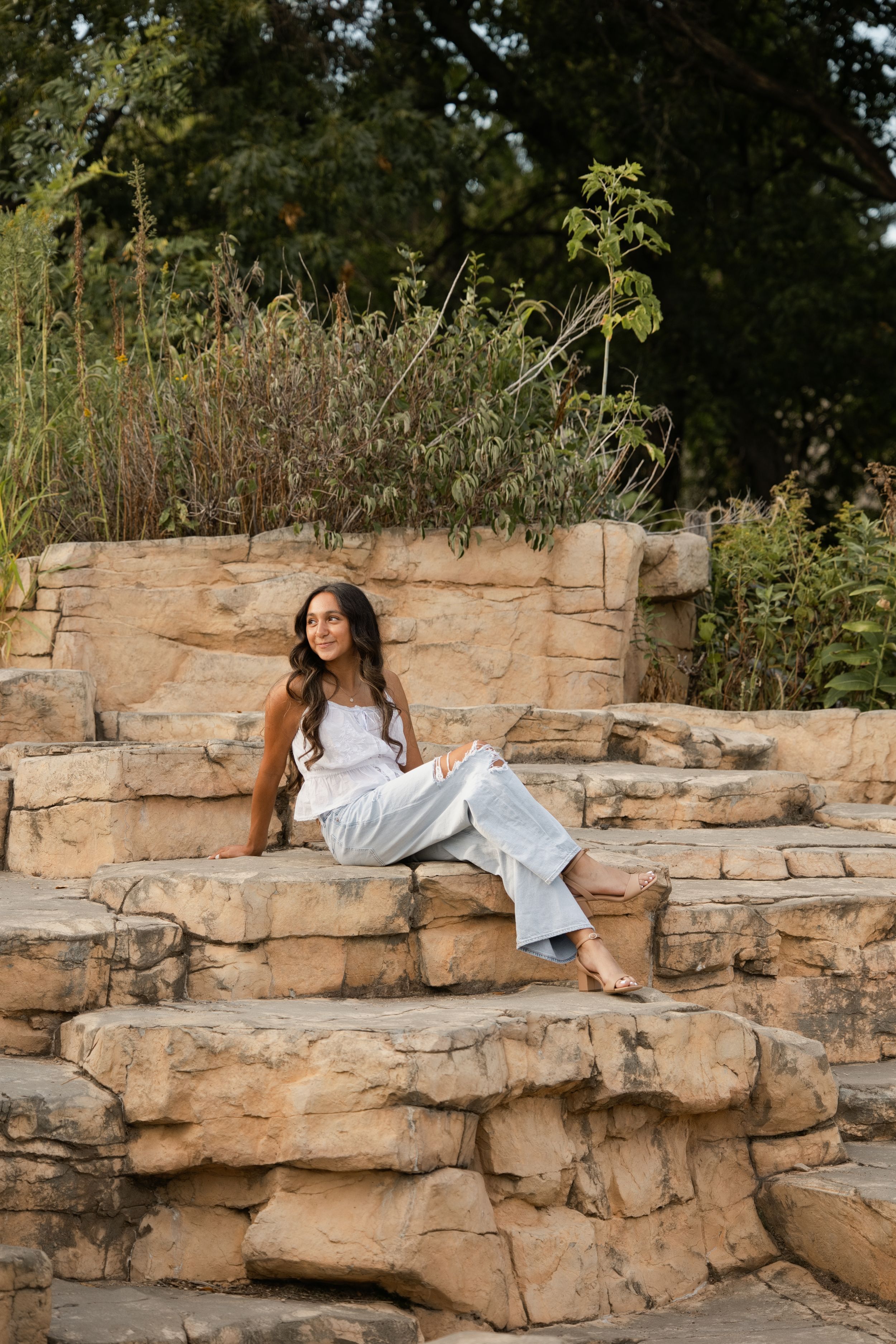 Young woman in white top and light pants sitting on natural stone steps surrounded by lush greenery outdoors.