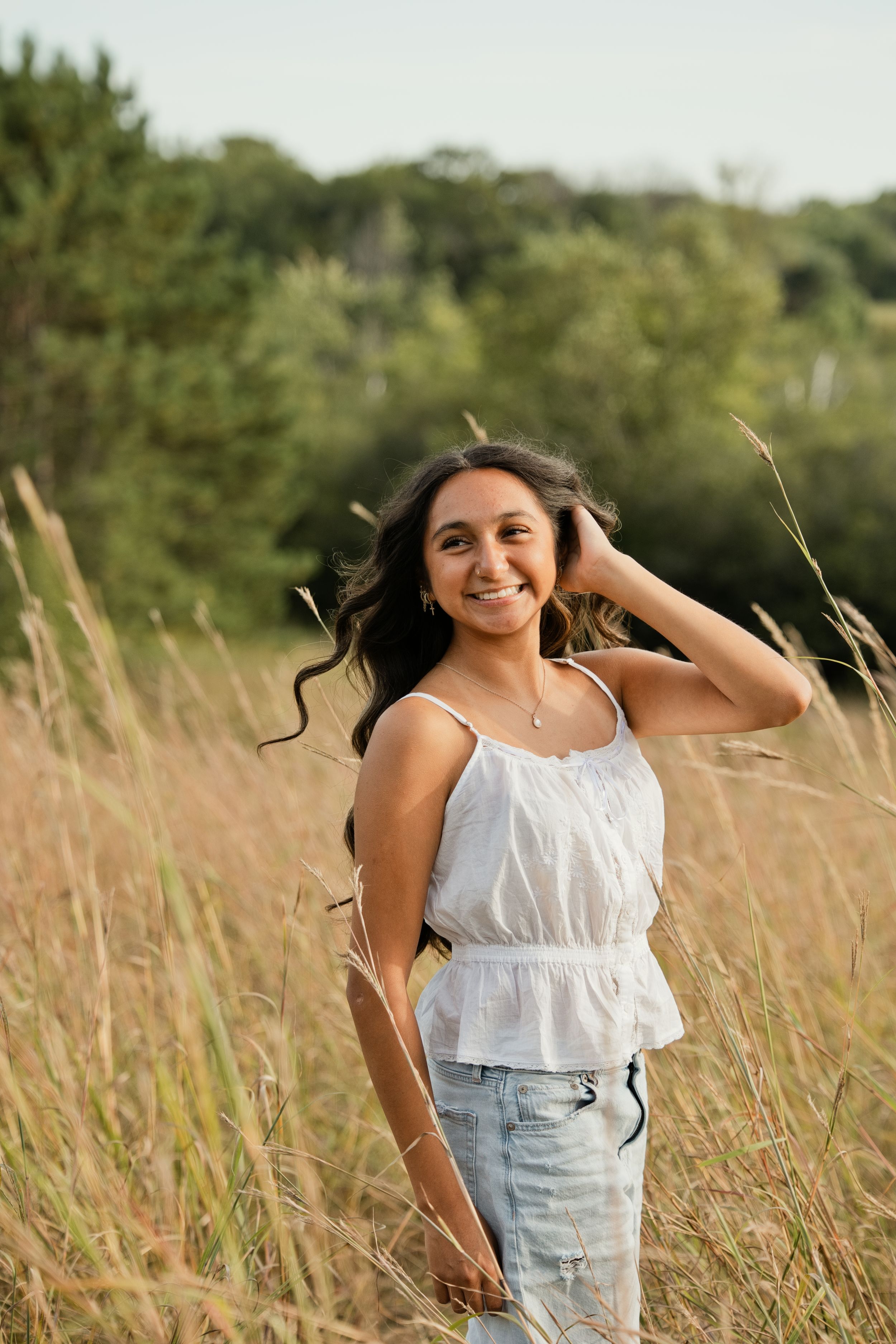Young woman in white top and jeans smiling in golden field with green trees in background.