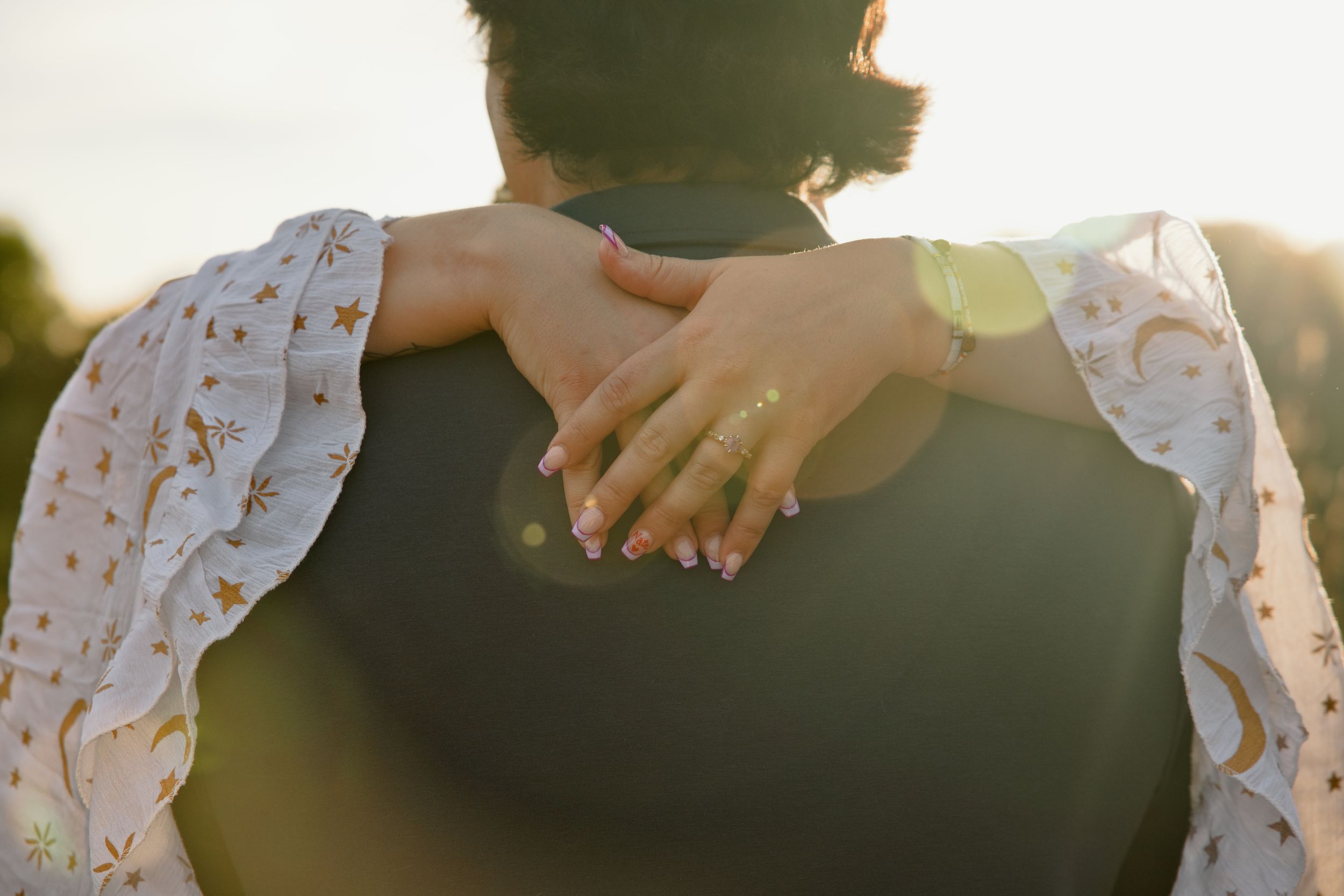 Couple embracing outdoors at golden hour, woman in floral dress with manicured nails resting hand on partner's back.