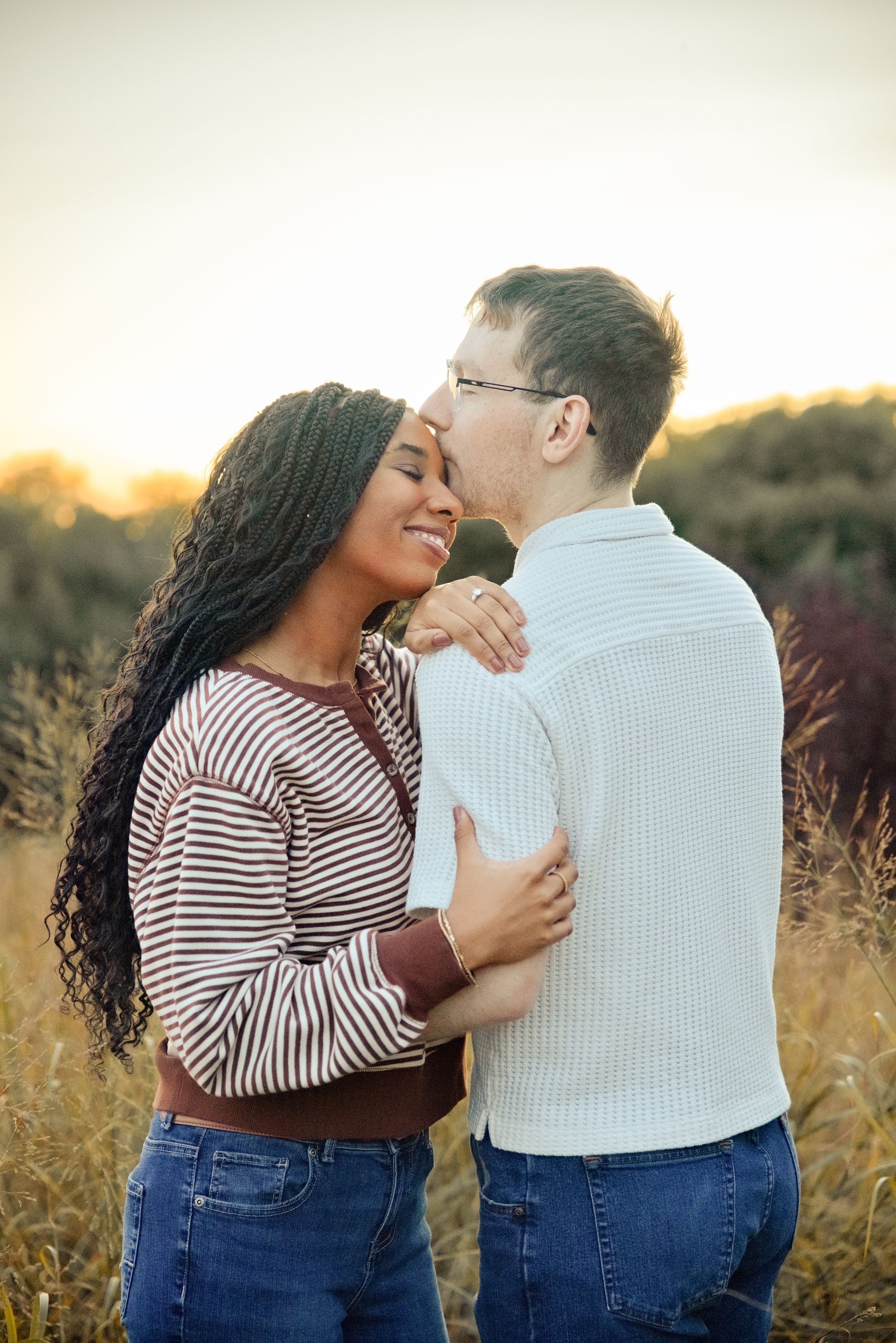 Interracial couple embracing and kissing in a golden field at sunset, woman smiling in striped top and jeans.