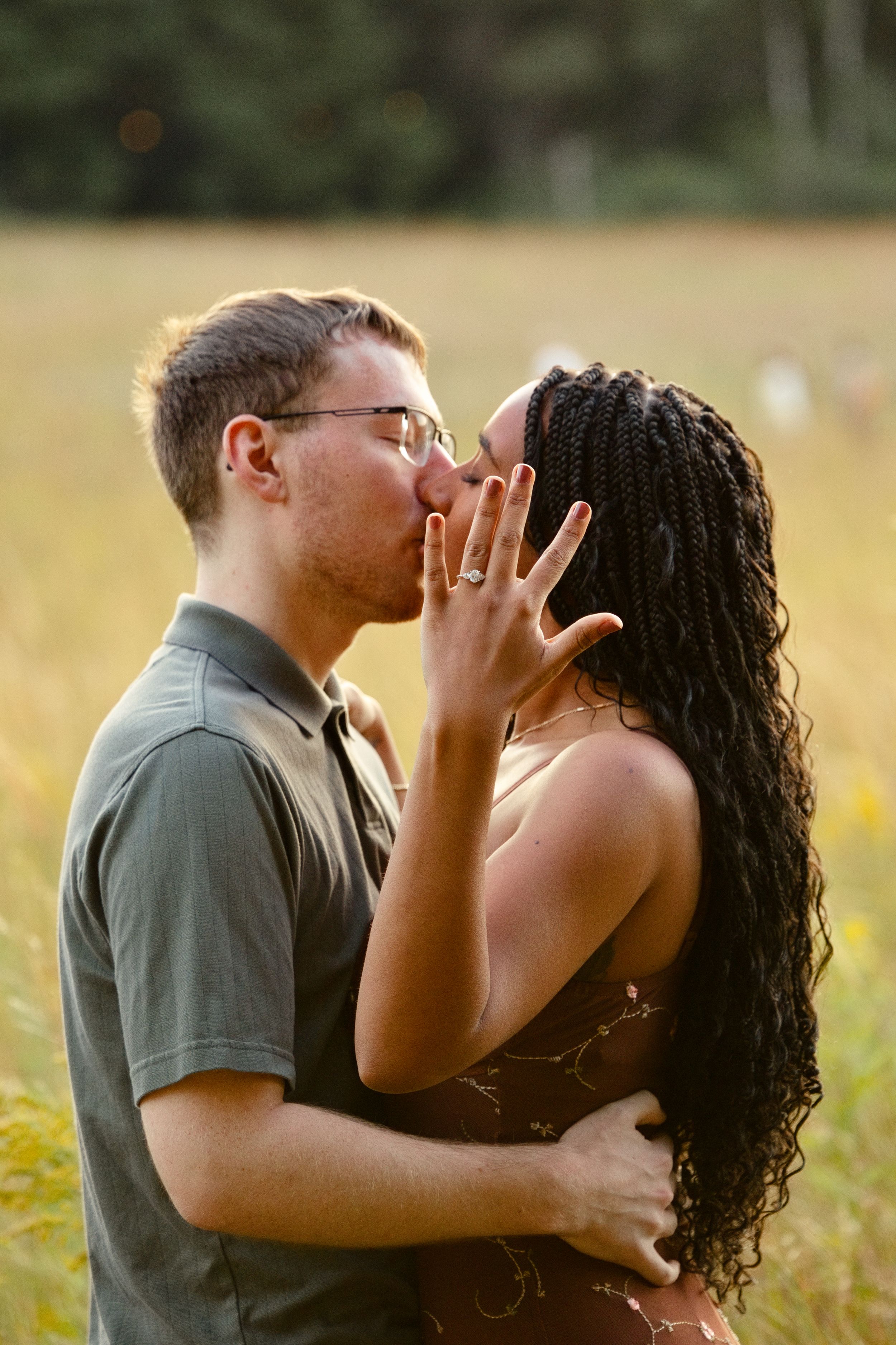 Couple sharing a romantic kiss in a golden field, woman showing off engagement ring with braided hair.