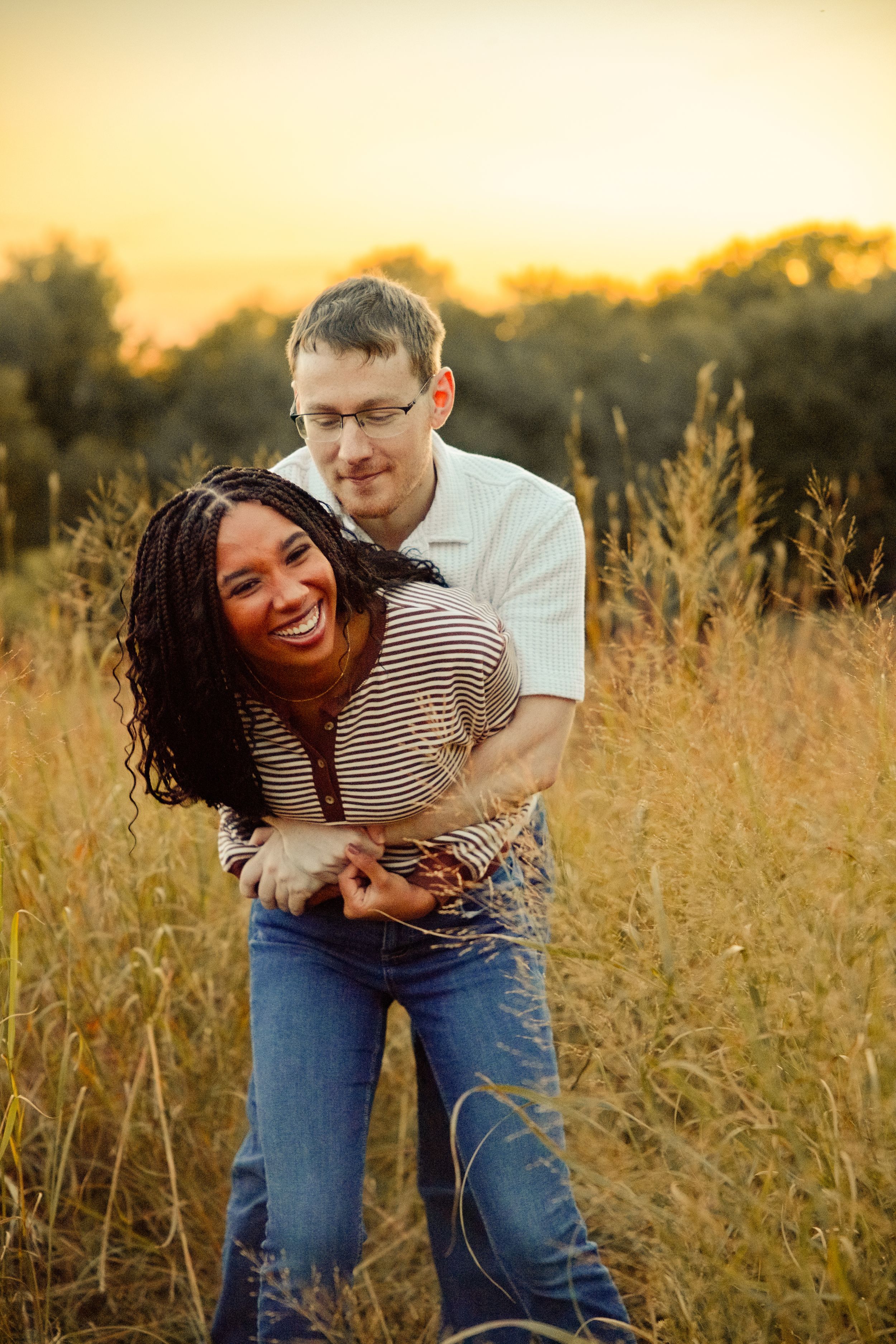 A joyful interracial couple embracing in a golden field at sunset, laughing together in casual attire.