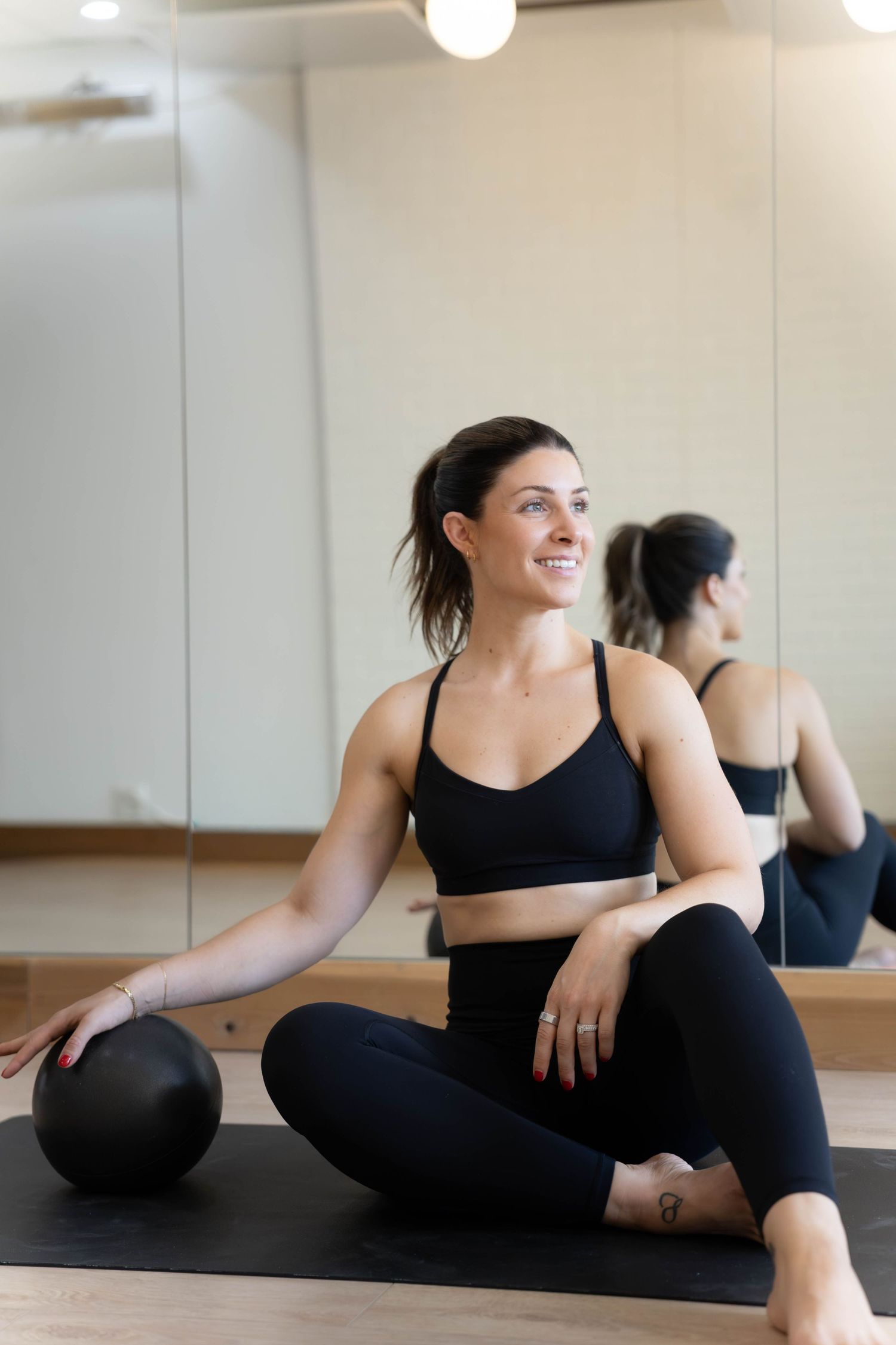 Smiling woman in black activewear sitting on yoga mat with pilates ball in a mirrored fitness studio.