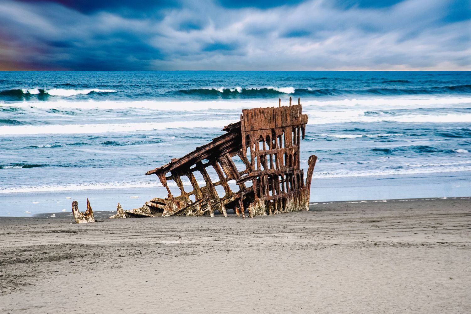 Rusty shipwreck remains on a sandy ocean beach with dramatic stormy blue sky and crashing waves in the background.