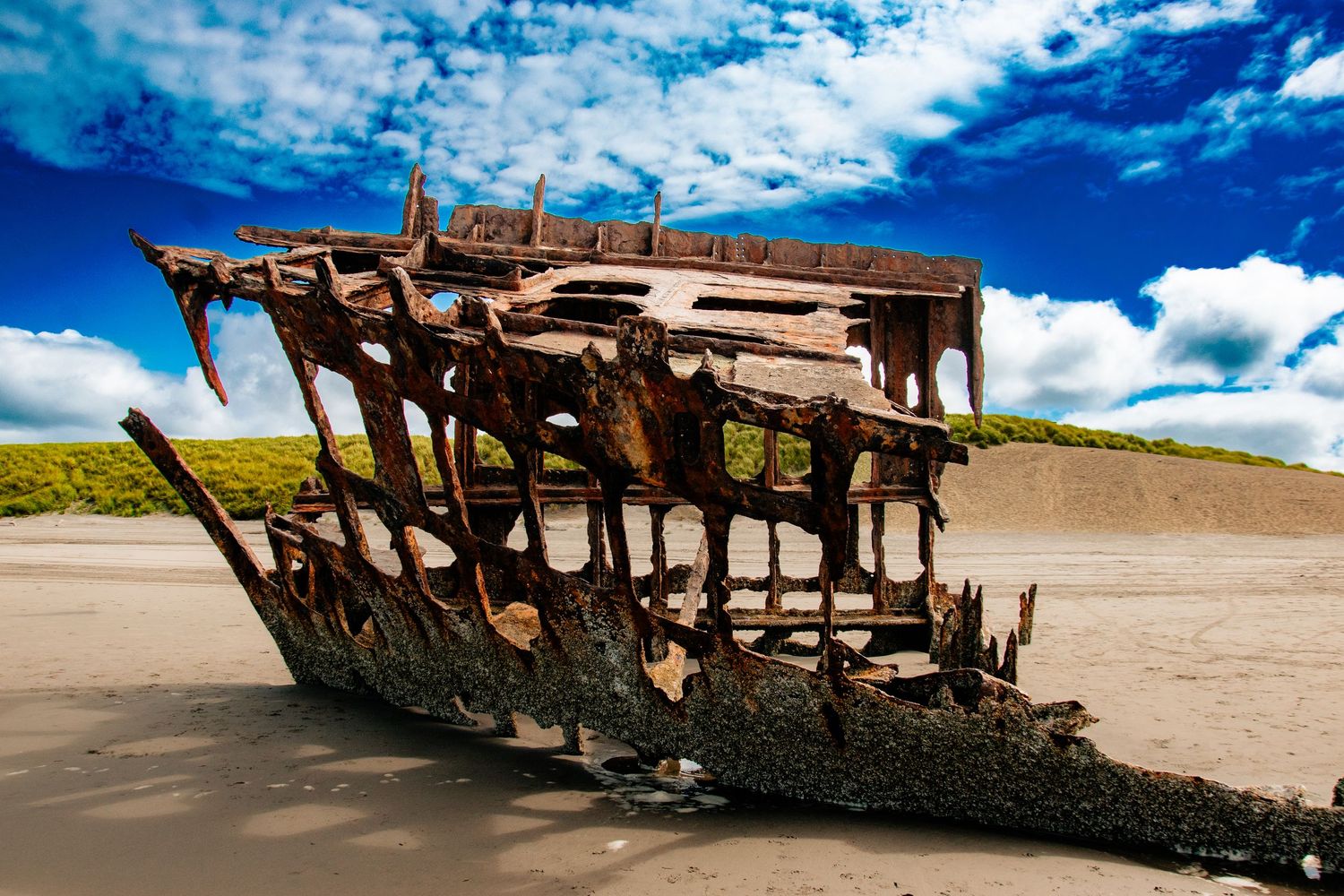 Rustic wooden shipwreck on sandy beach under dramatic blue sky with clouds.