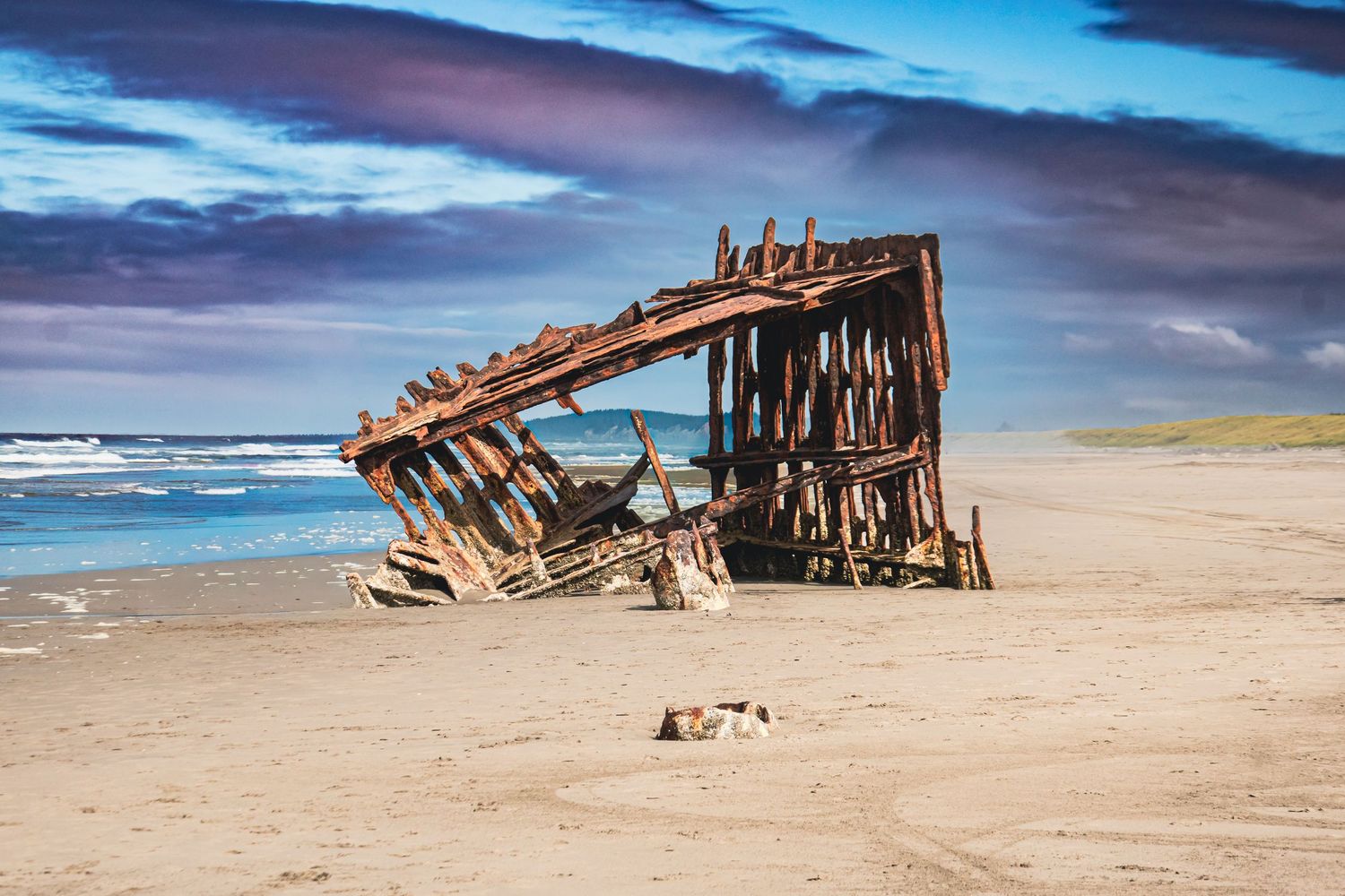 Weathered rusty shipwreck sitting on sandy beach with dramatic cloudy sky and ocean waves behind it.