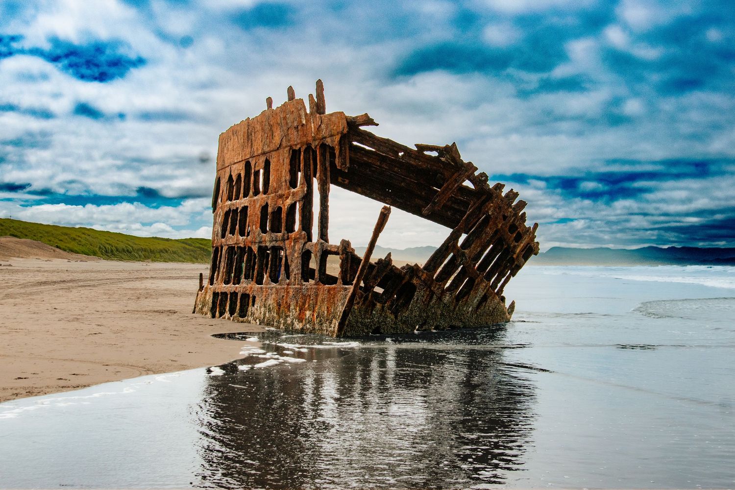 Rusty shipwreck remains on sandy beach with dramatic cloudy sky reflected in shallow water.
