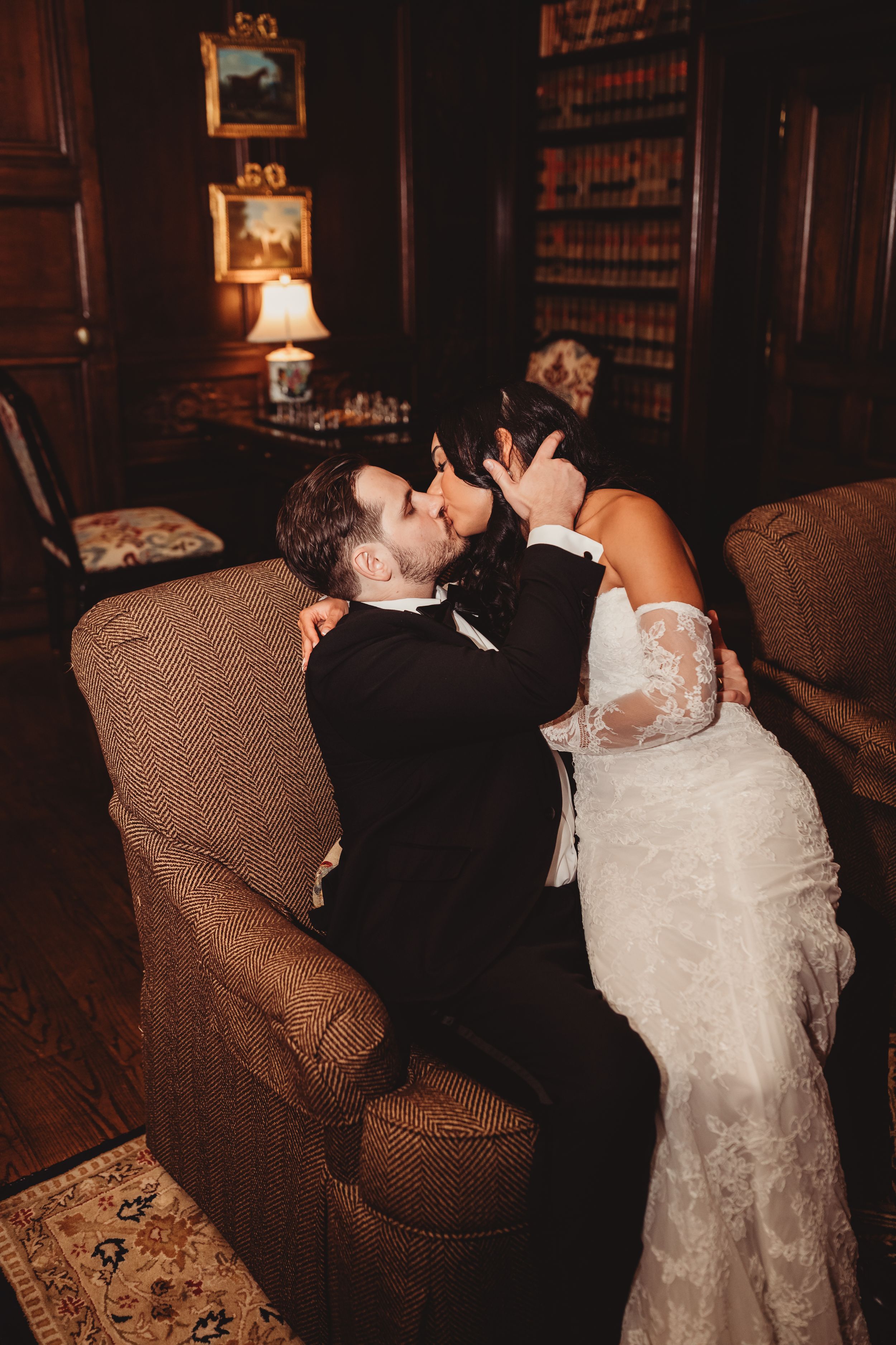 A bride and groom share a romantic kiss while seated on a brown armchair in an elegant wood-paneled library setting.