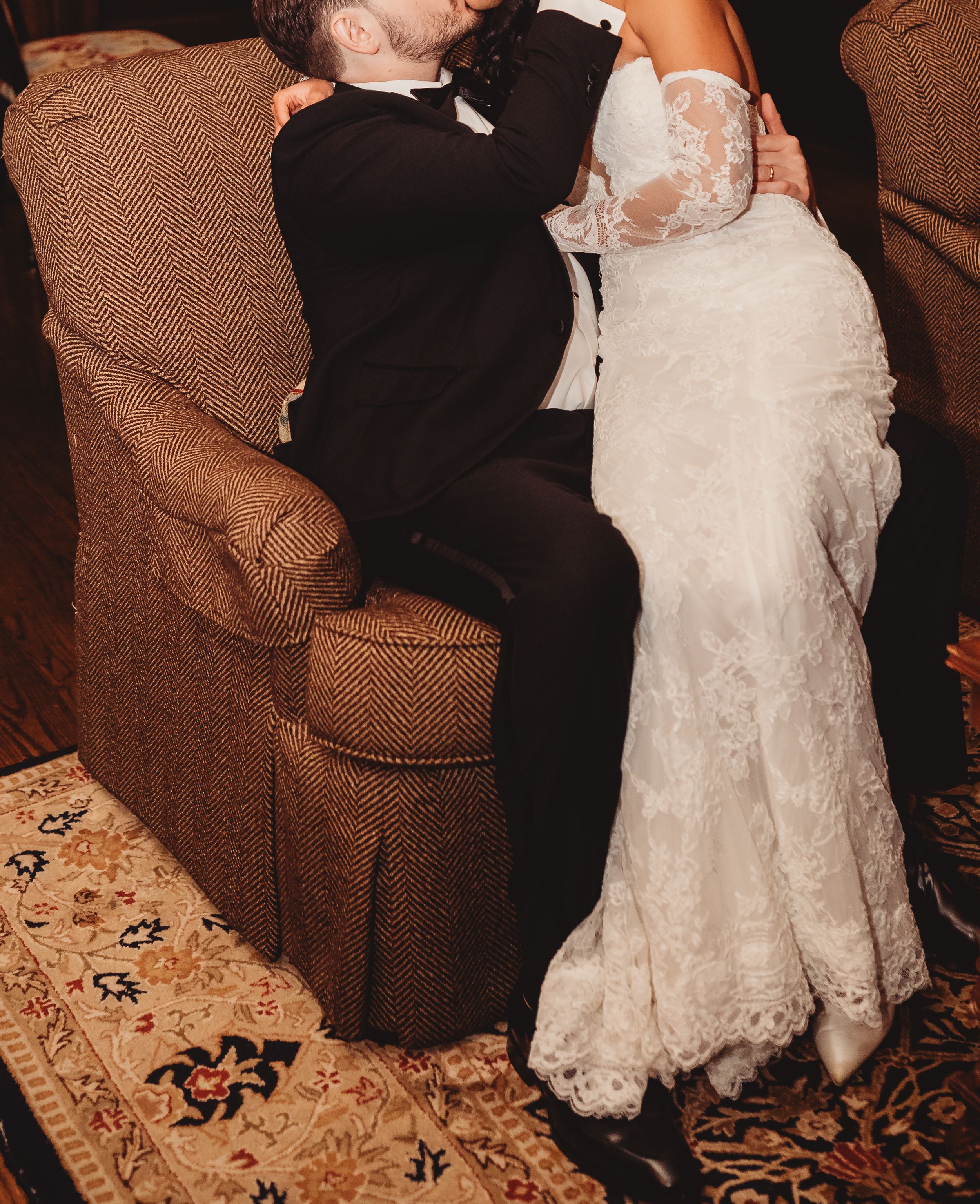 Bride and groom sharing a romantic kiss on a brown armchair, bride in lace wedding gown, groom in black tuxedo.