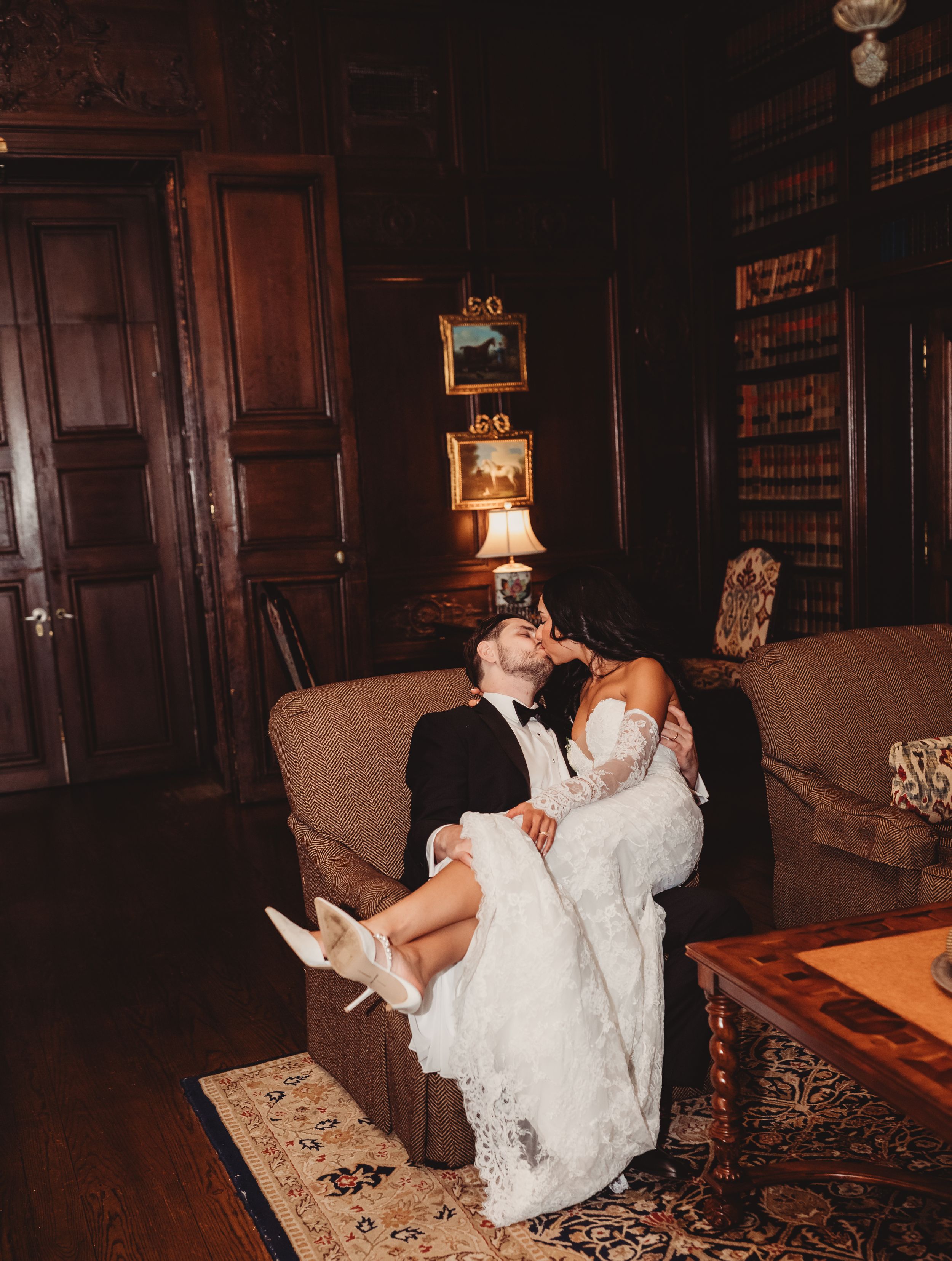 Bride and groom share a romantic kiss on a sofa in an elegant wood-paneled library during their wedding day.