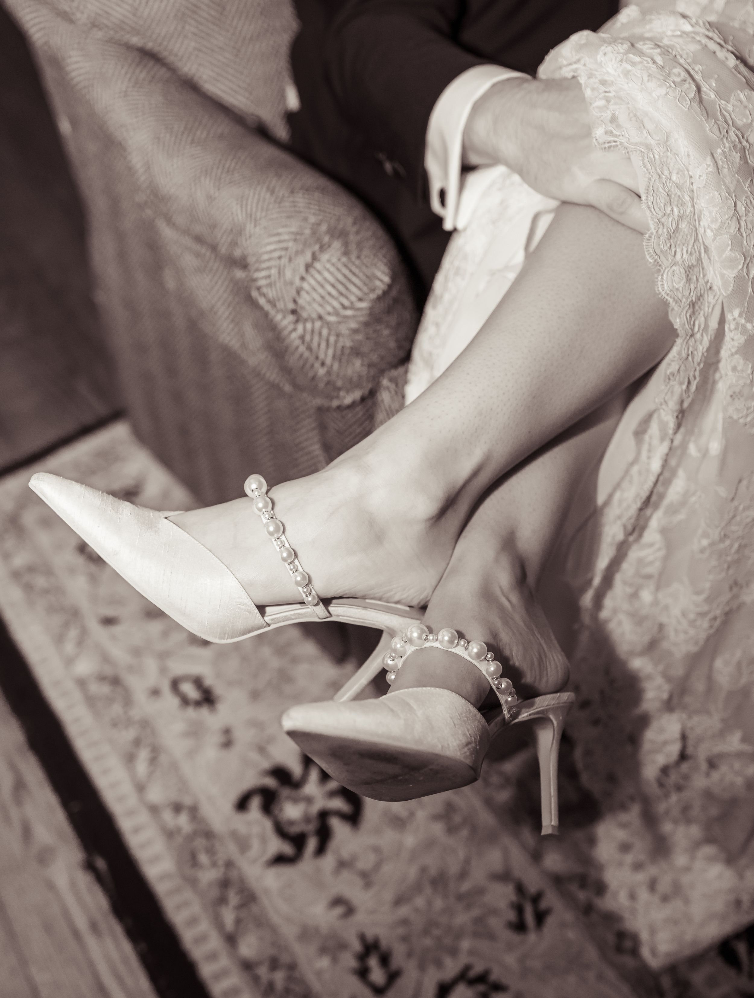 Elegant bridal heels with pearl ankle strap details, worn by a bride in a lace wedding dress, sepia tone.