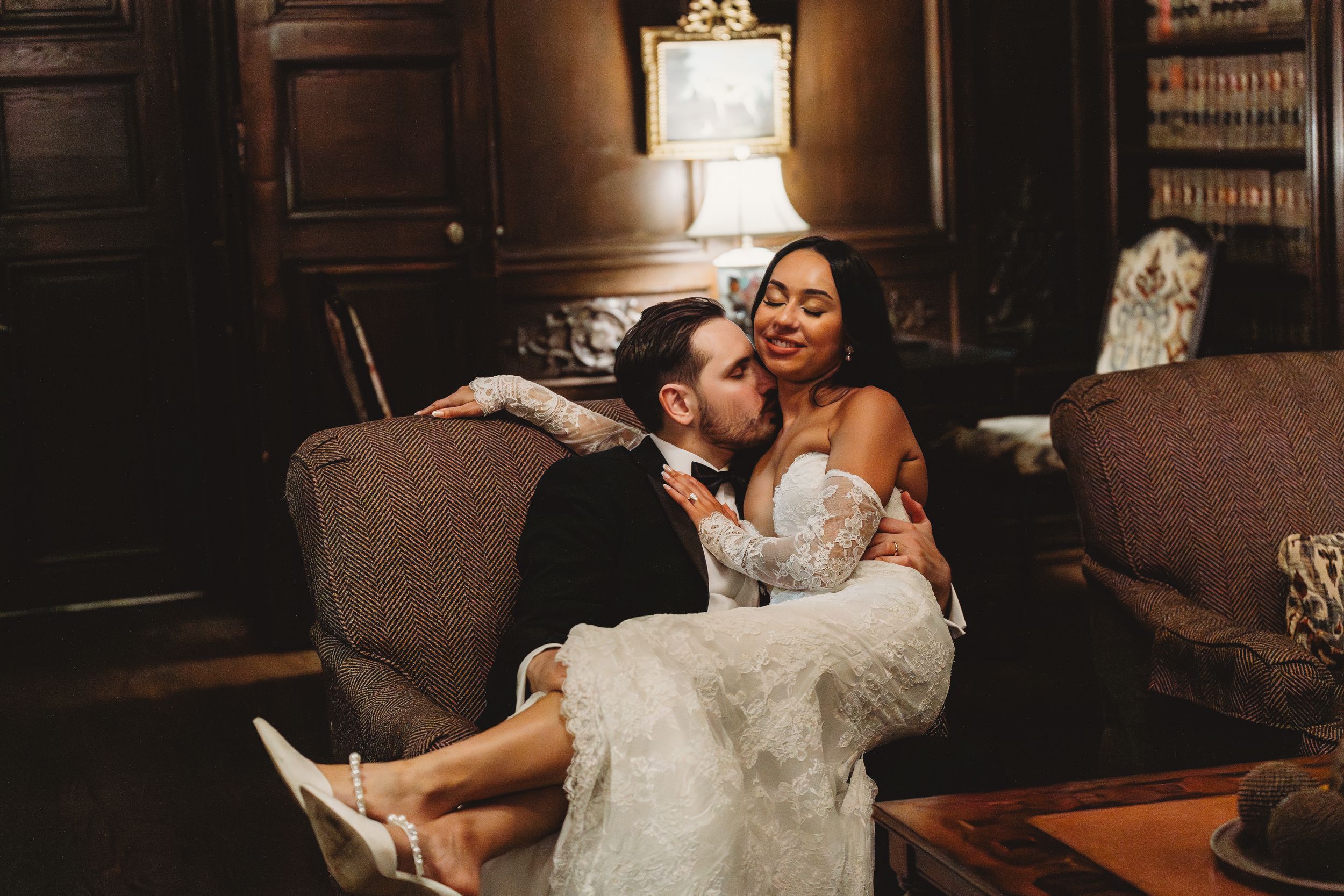 A bride and groom share an intimate moment on a vintage sofa in an elegant library setting during their wedding day.