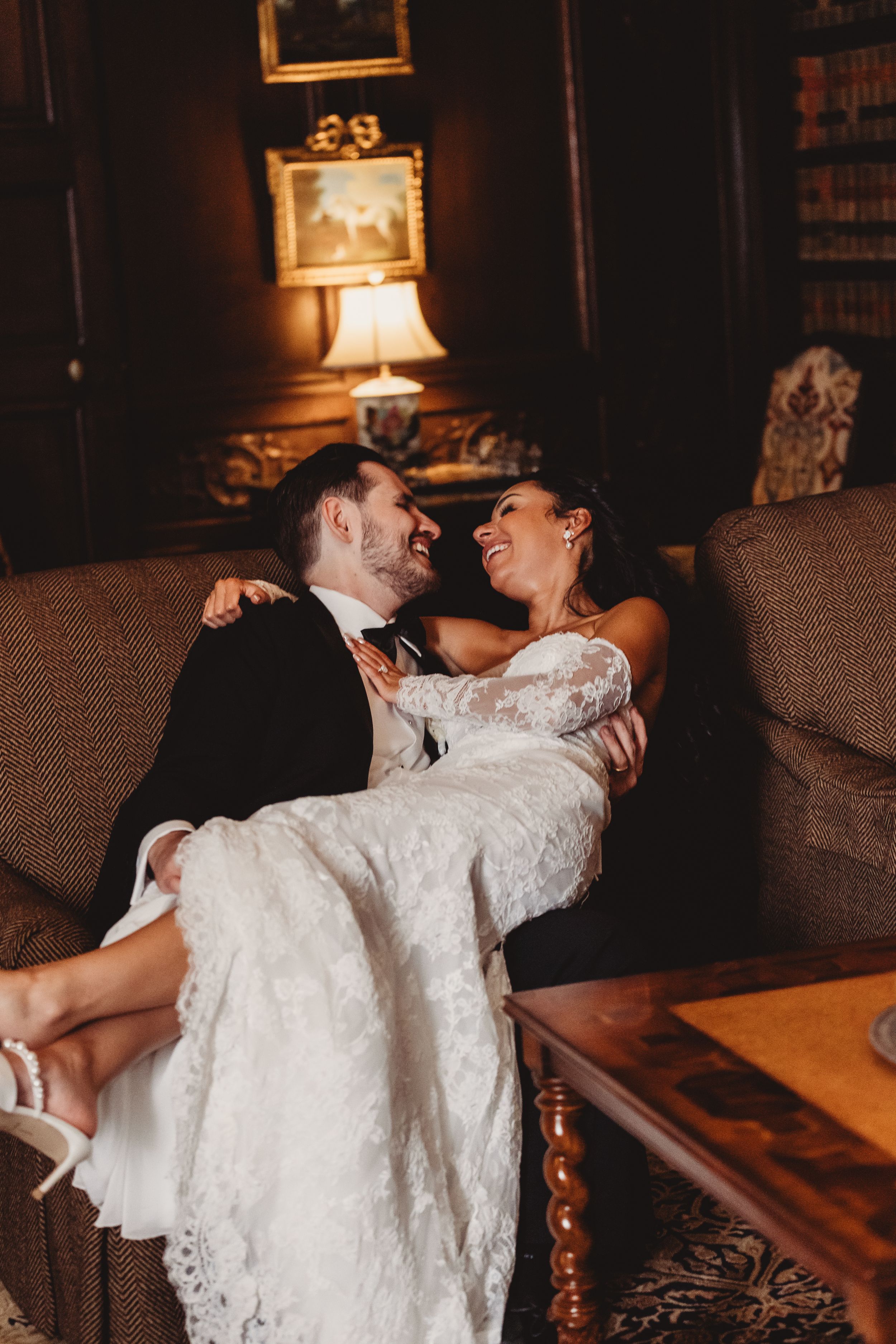 A bride and groom share a joyful moment on a couch, with the groom holding the bride in his arms in an elegant room.