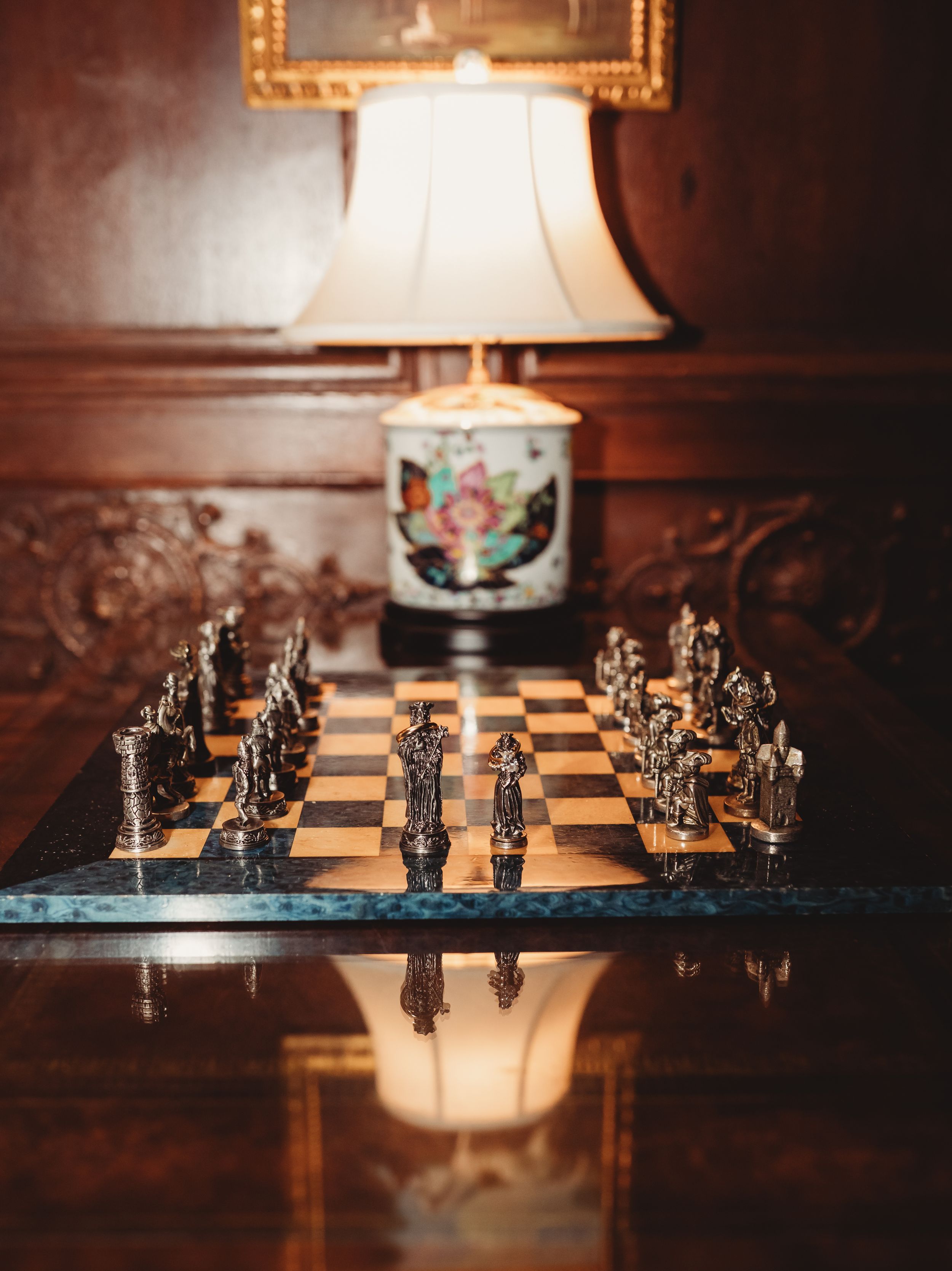 Ornate chess set with metallic pieces on a marble board atop a wooden table, with a floral lamp in the background.