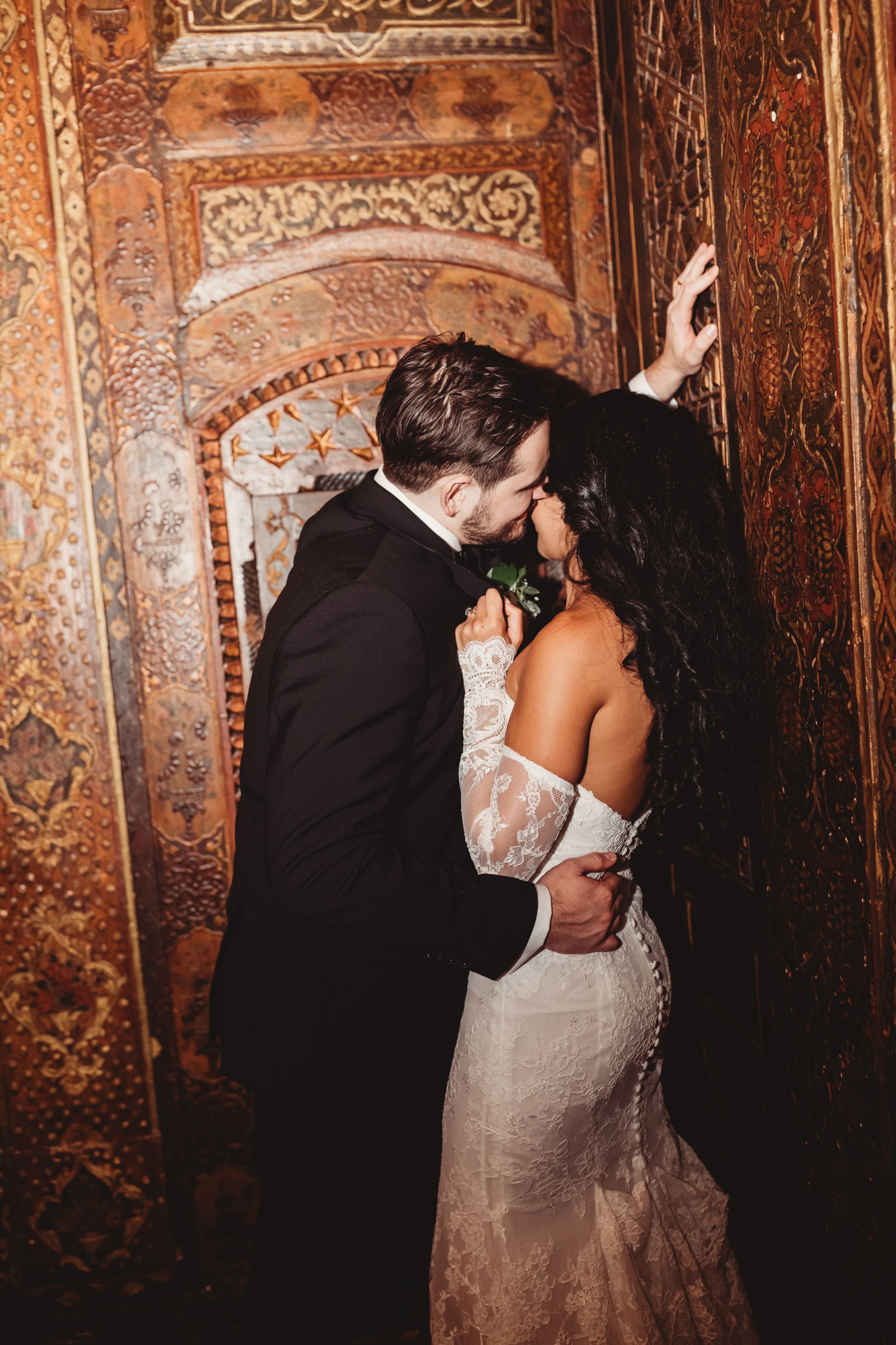 Bride and groom share a romantic kiss against ornate golden carved doors in wedding attire.