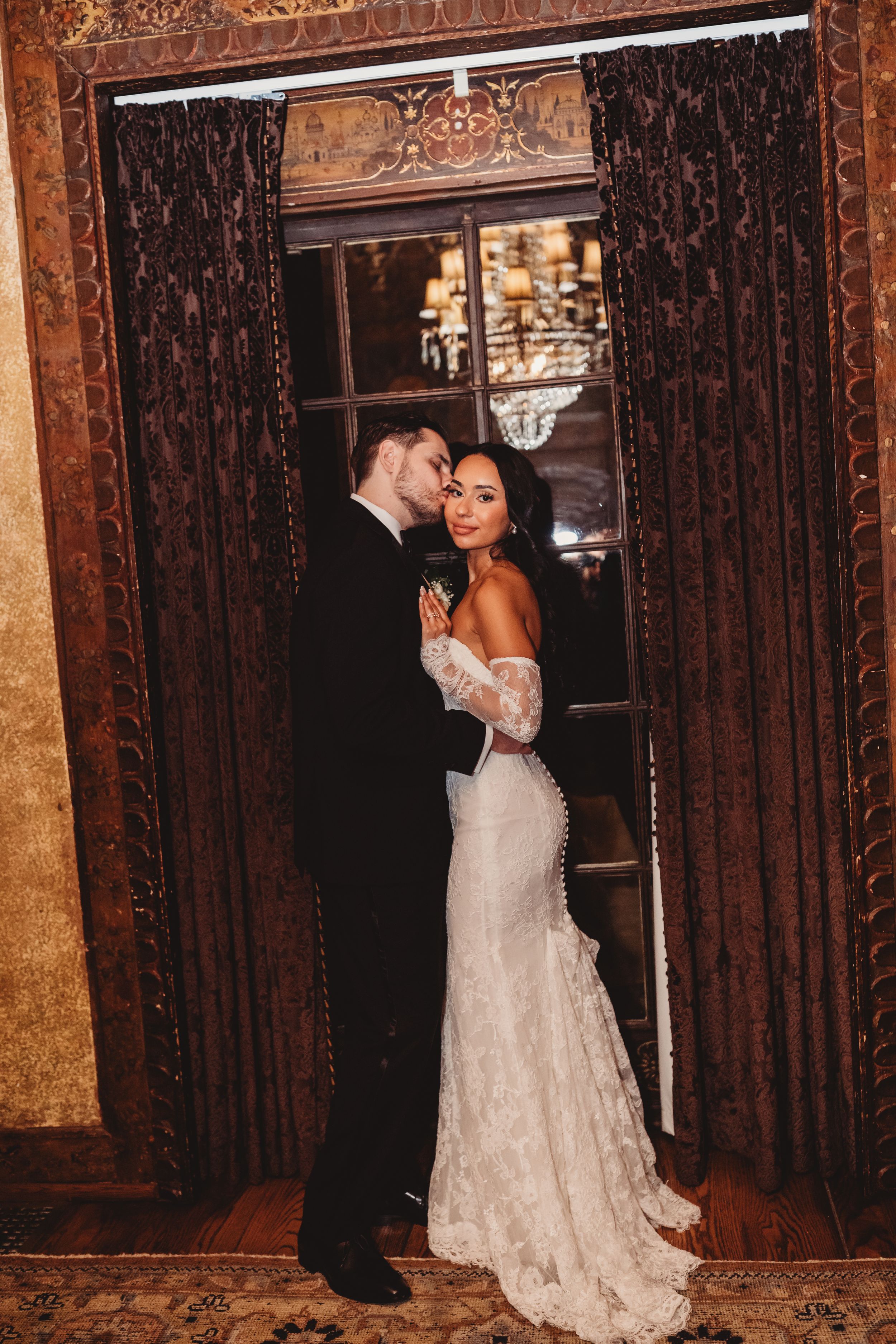 Bride in lace gown and groom in black tuxedo share romantic embrace by ornate curtained window in elegant venue.