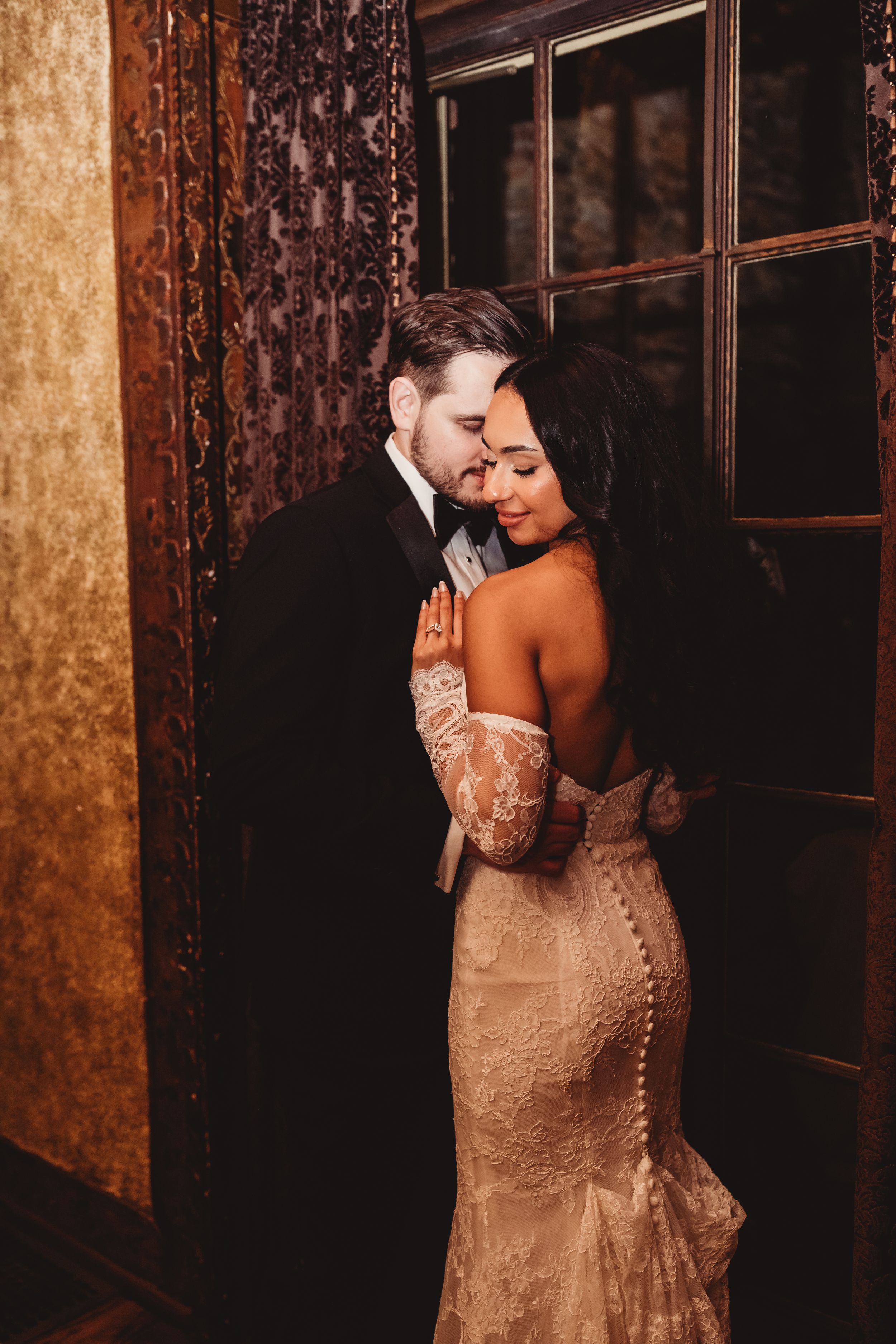 Groom in black tuxedo embraces bride in lace off-shoulder gown near decorative golden wall.